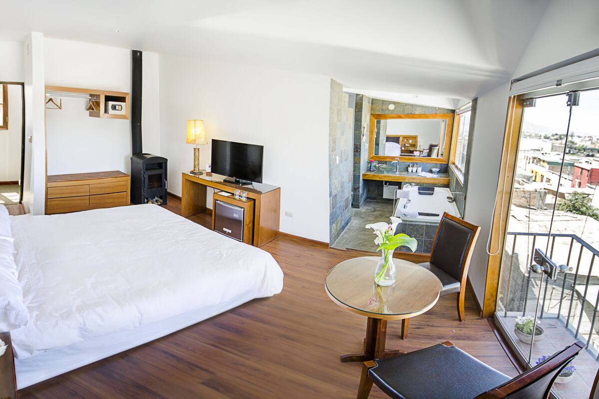 Habitación de hotel con cama, escritorio, televisión, balcón con vistas y baño con espejo y lavamanos.