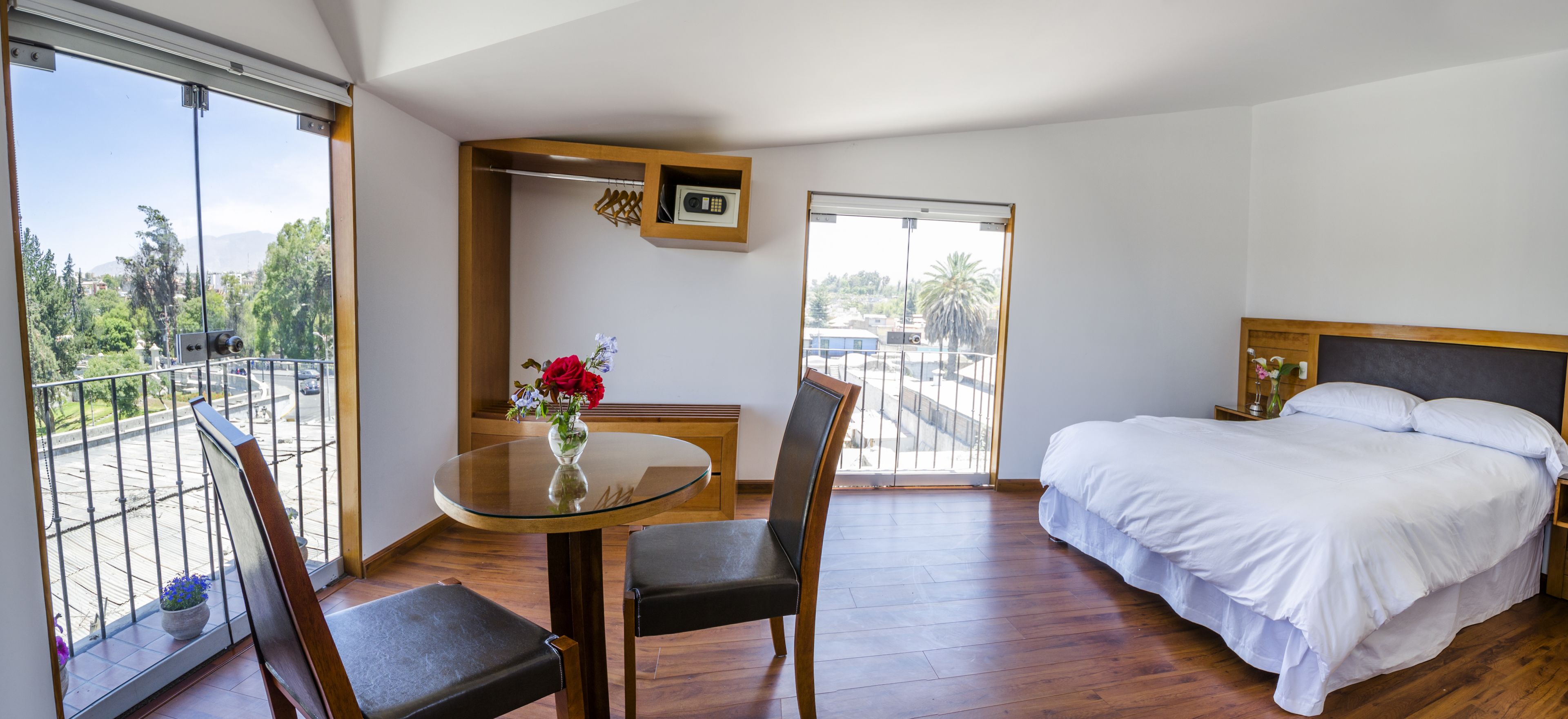Habitación de hotel con balcón, una cama, una mesa redonda con sillas y un estante pequeño con un teléfono y objetos decorativos.