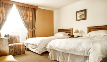 Habitación de hotel con dos camas, una mesita de noche con lámpara, una silla y un cuadro en la pared.