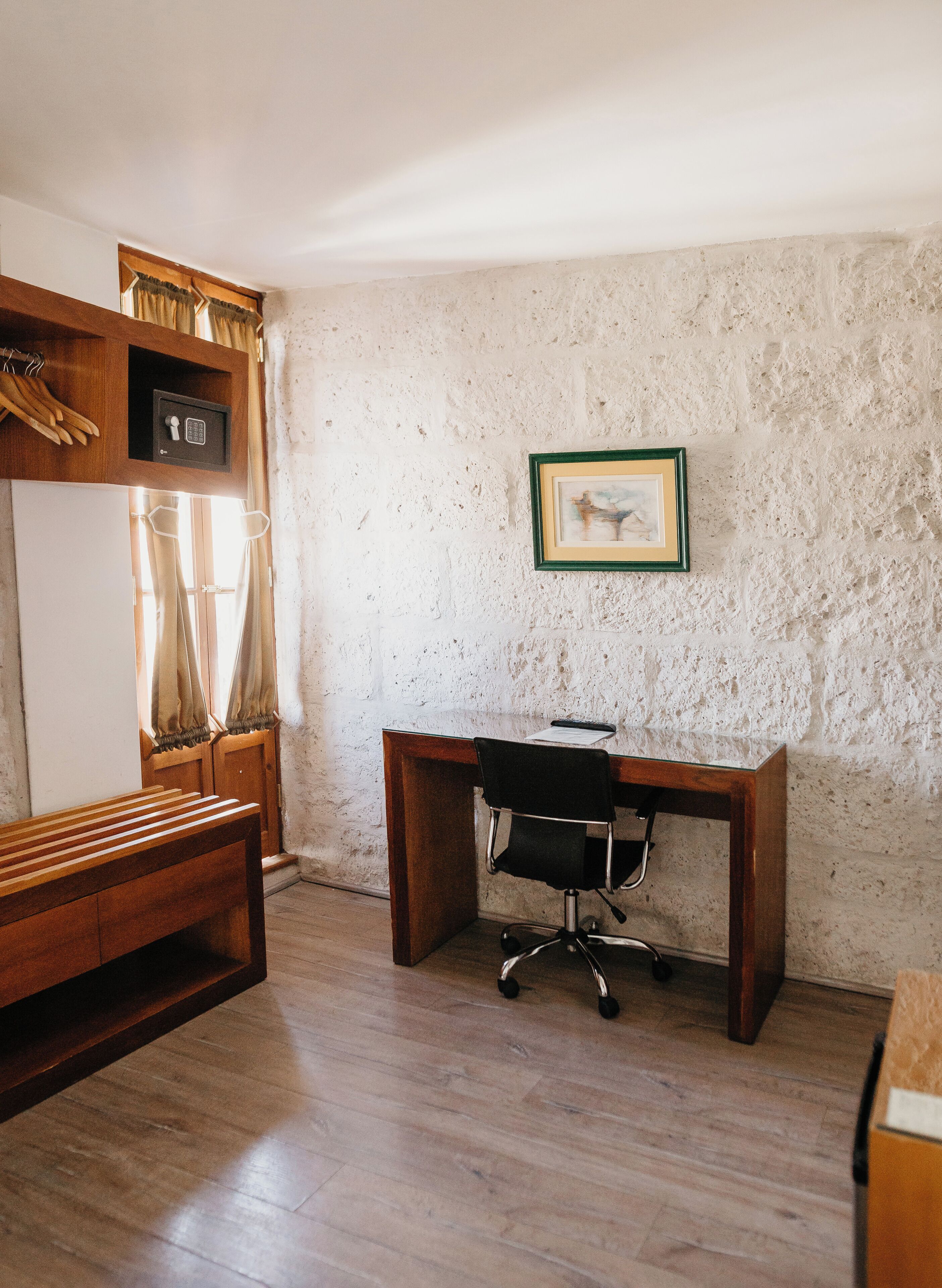 A room with a wooden desk, a chair, a small safe, a framed picture on the wall, and a bench.