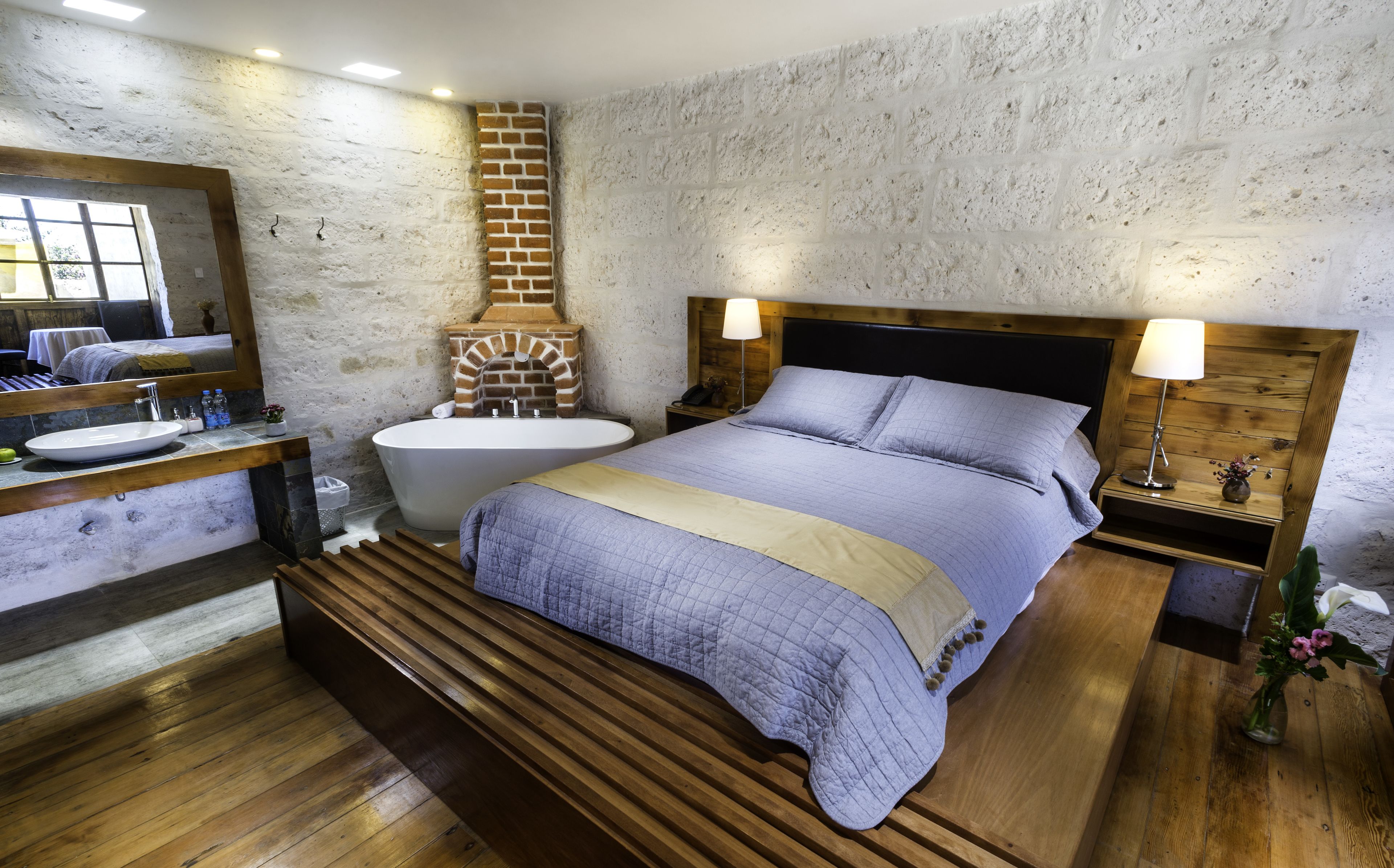 Quarto com cama grande, cabeceira de madeira, mesa de cabeceira, espelho e banheiro com pia, vaso sanitário e lareira de tijolos.