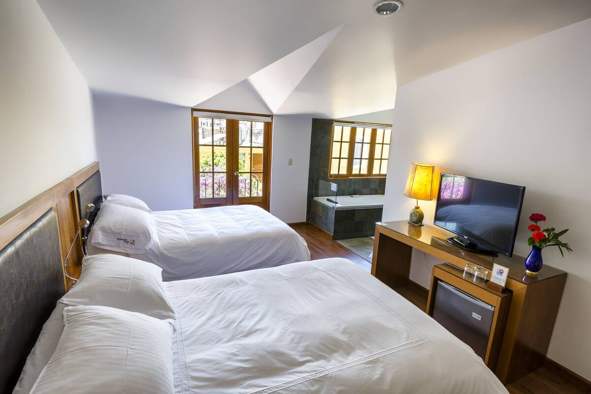 Habitación de hotel con dos camas, televisor, escritorio con lámpara y refrigerador pequeño.