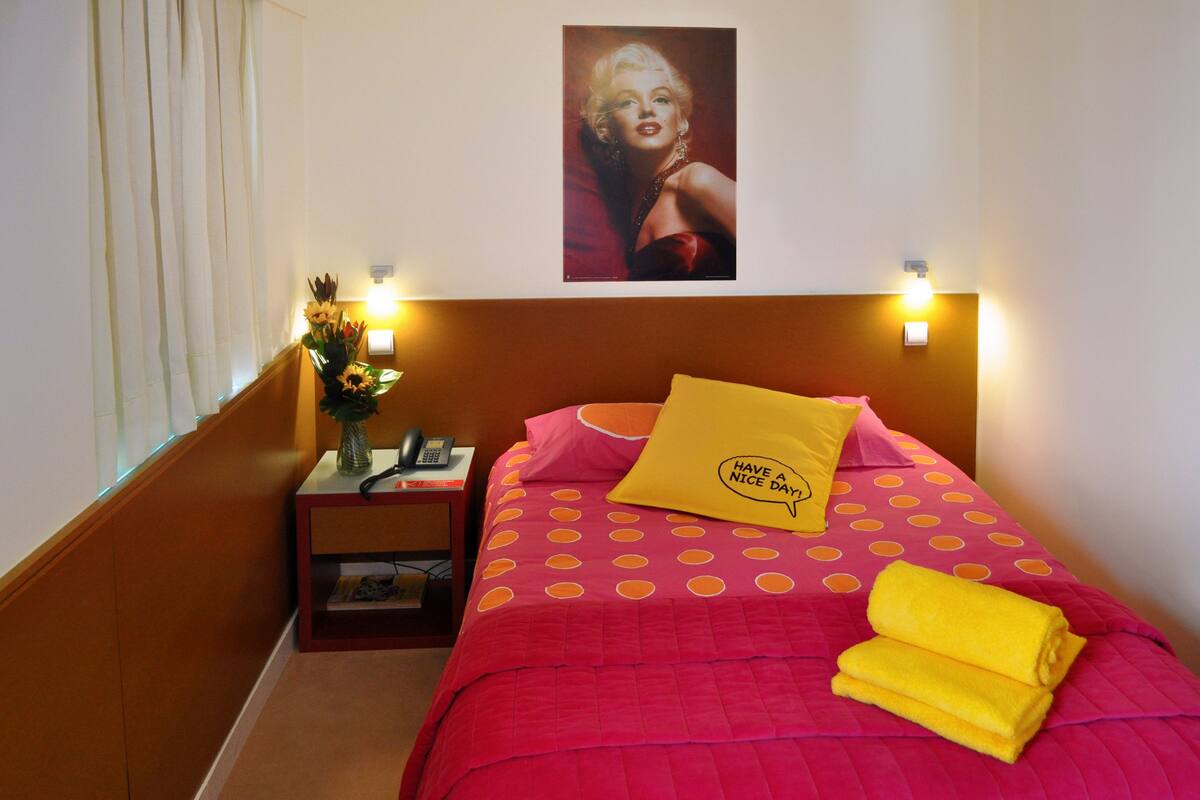 Habitación de hotel con una cama que tiene una colcha de lunares, un almohadón amarillo con un mensaje y un cuadro en la pared.