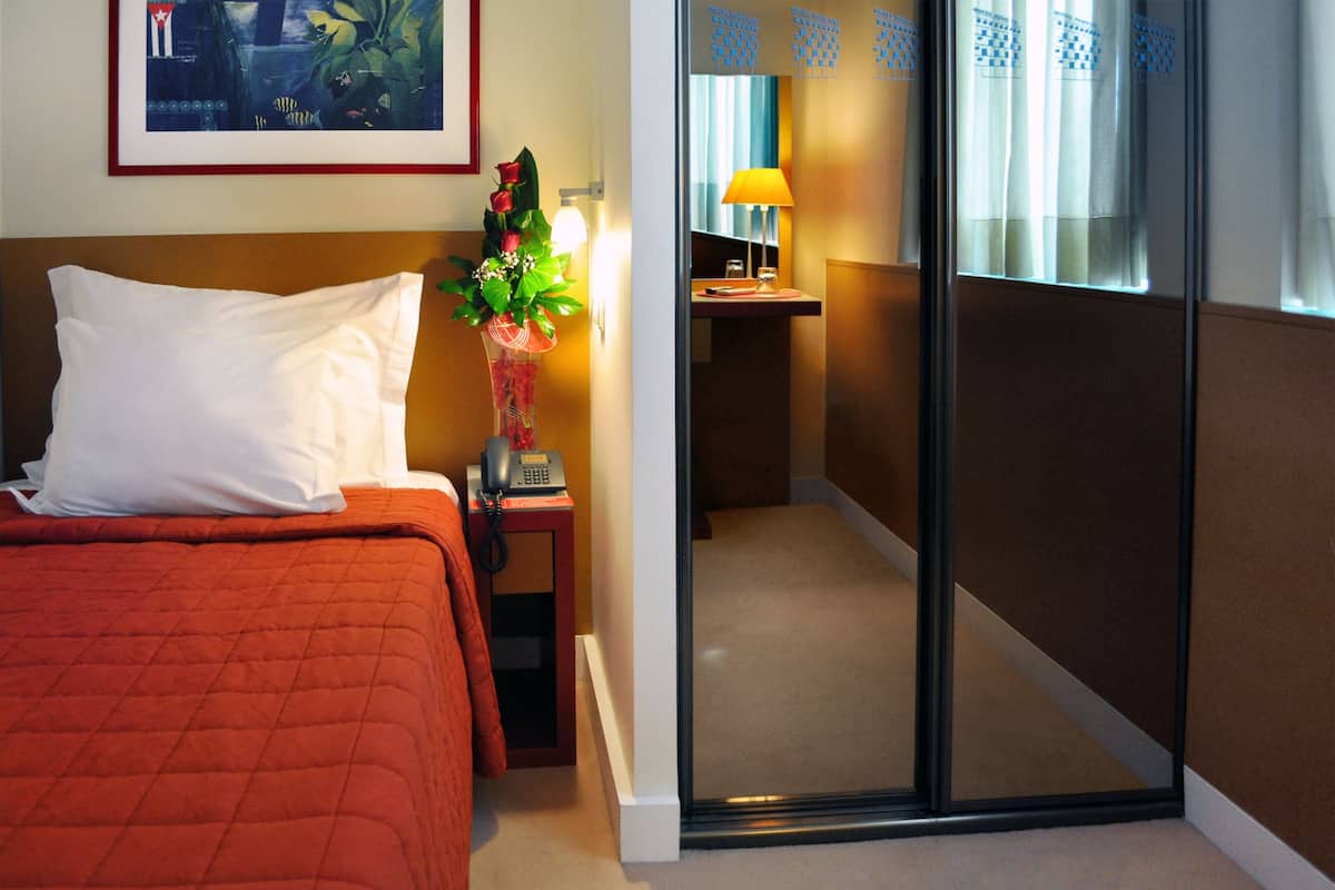 Una habitación de hotel con una cama, una mesita de noche con lámpara, un florero con flores y una puerta corredera que da a un baño.