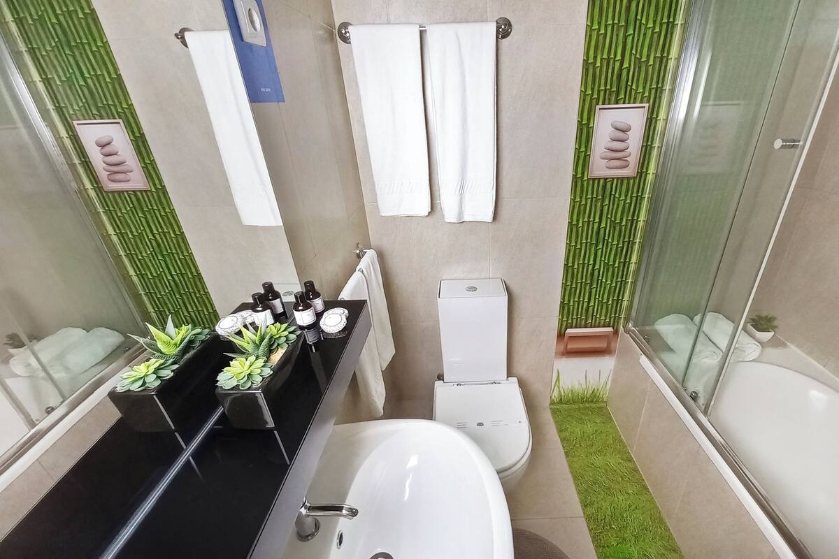 Un baño moderno con tocador negro, lavamanos blanco y retrete. Hay dos toallas blancas colgadas en la pared y una pequeña planta en maceta sobre el tocador.