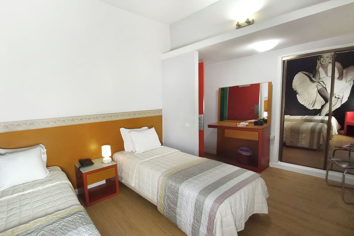 Una habitación de hotel con dos camas individuales, una mesita de noche pequeña con lámpara, un escritorio con silla y un espejo grande.