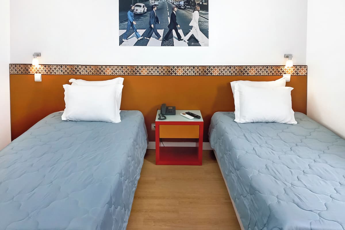 Una habitación de hotel con dos camas individuales, una mesita con teléfono y control remoto, y un cuadro de The Beatles en la pared.