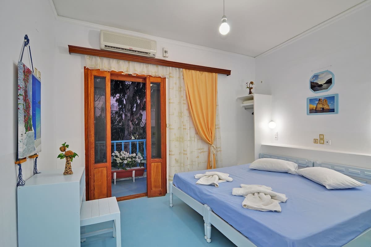 A bedroom with a bed, a bedside table, a small desk, a potted plant, and a window with curtains.