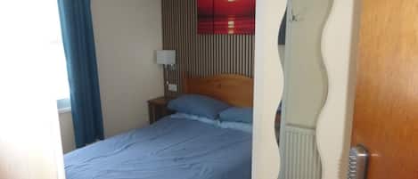 A bedroom with a bed, a nightstand, a wall-mounted lamp, a mirror, and a radiator.