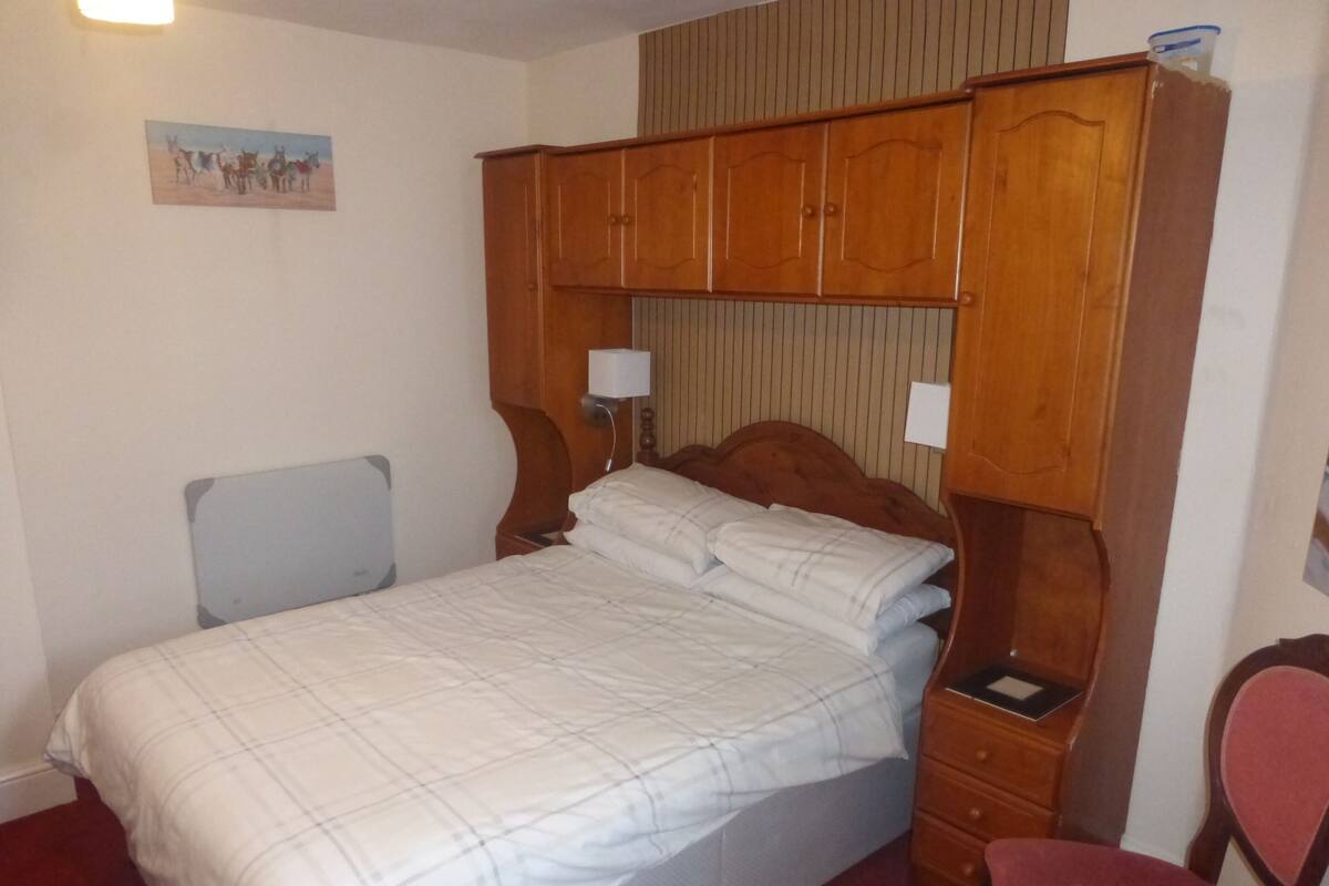 A bedroom with a bed, a wooden wardrobe, a bedside table with a lamp, a chair, and a wall-mounted picture.