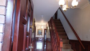 Una escalera de madera con barandilla que lleva a un piso superior. Un candelabro cuelga del techo, y se puede ver un cuadro enmarcado en la pared.