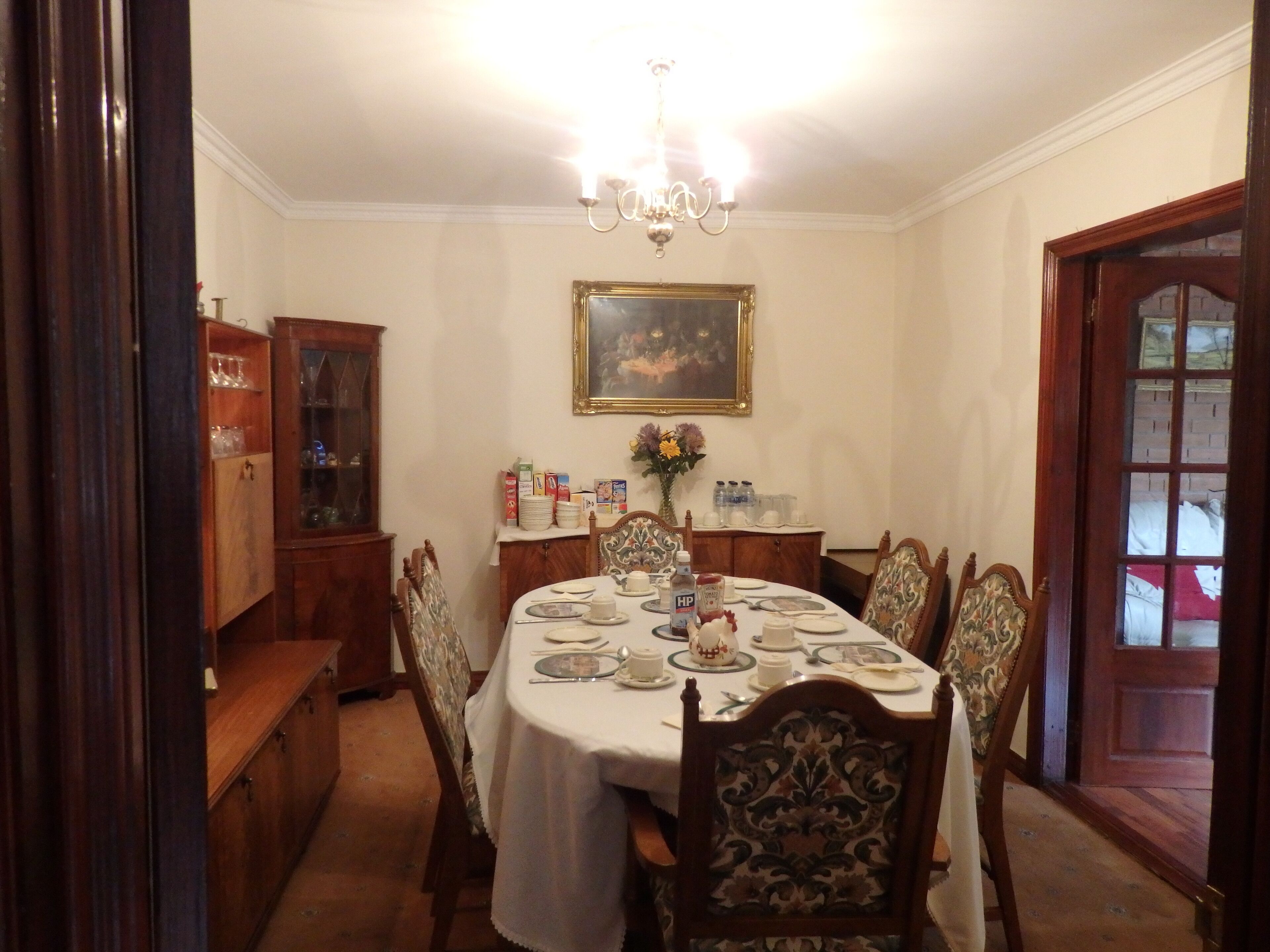 A dining room with a round table set for six, surrounded by ornate chairs. A chandelier hangs from the ceiling, and a framed painting is on the wall.