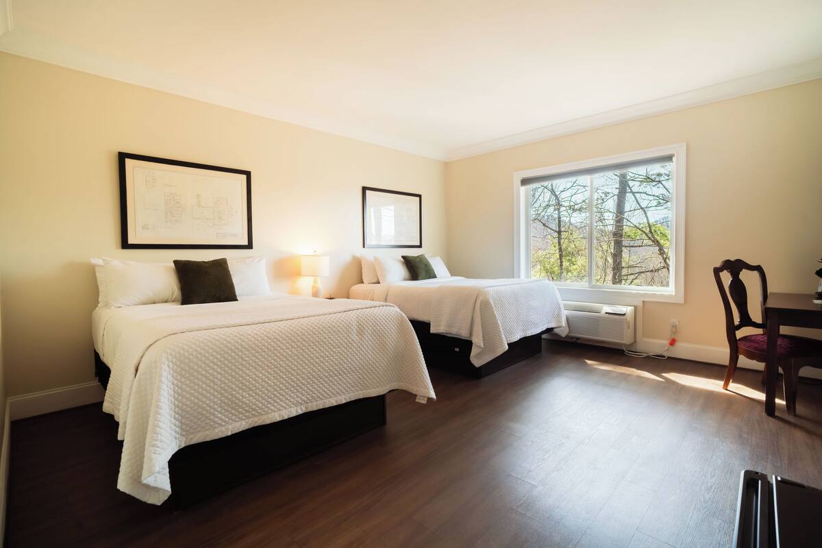 A hotel room with two beds, a large window, and a dining table.