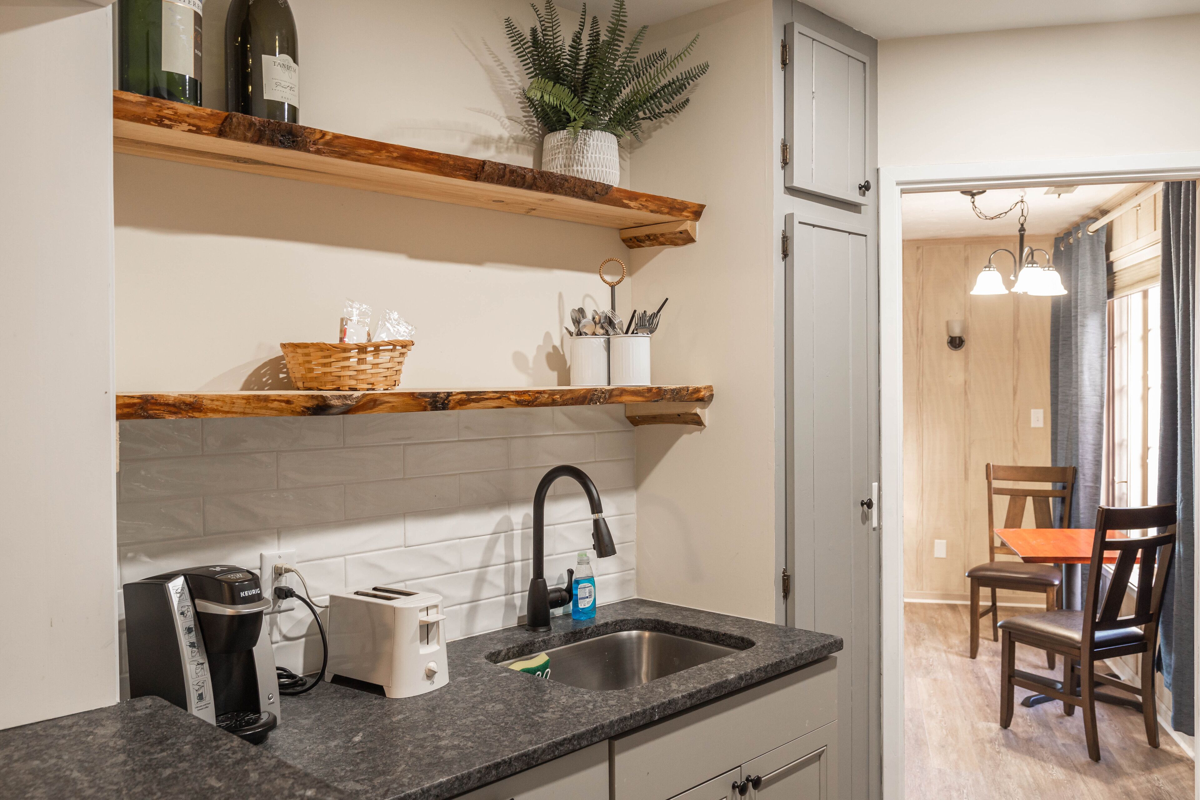 Una cocina con encimera de granito, cafetera, pileta y estanterías de madera.