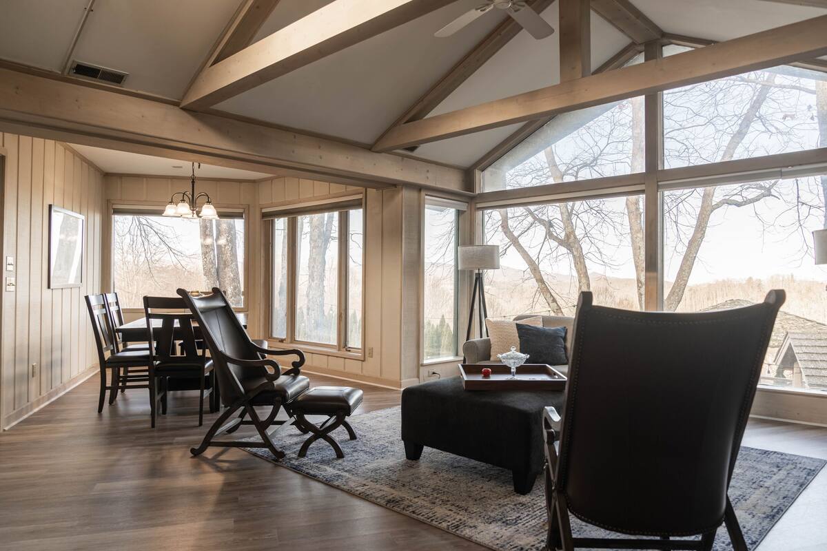 A spacious room with a dining area, a seating area with a sofa and chairs, large windows, and wooden beams on the ceiling.