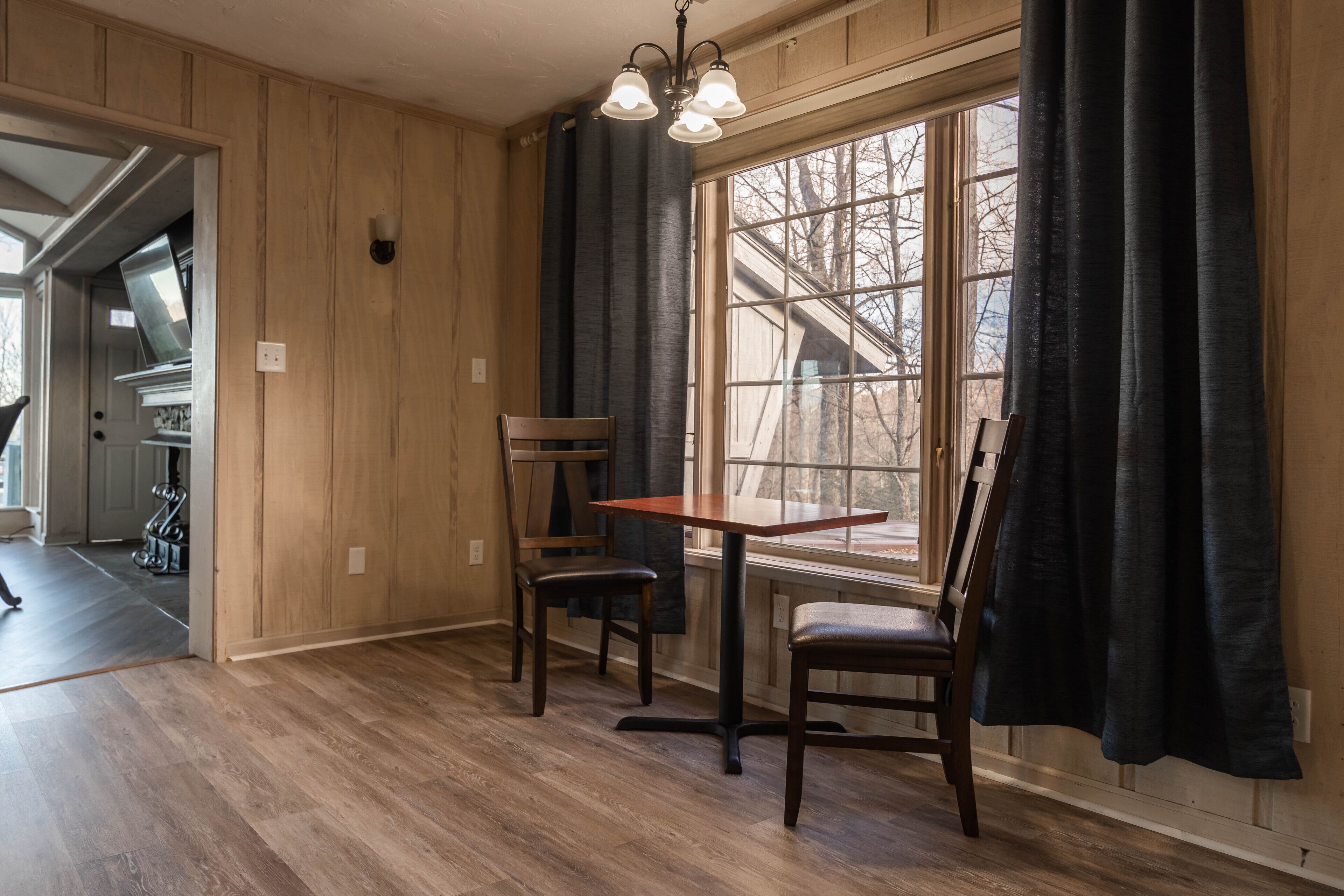 A room with wooden panel walls, a dining table with chairs, a window with curtains, and a ceiling light fixture.