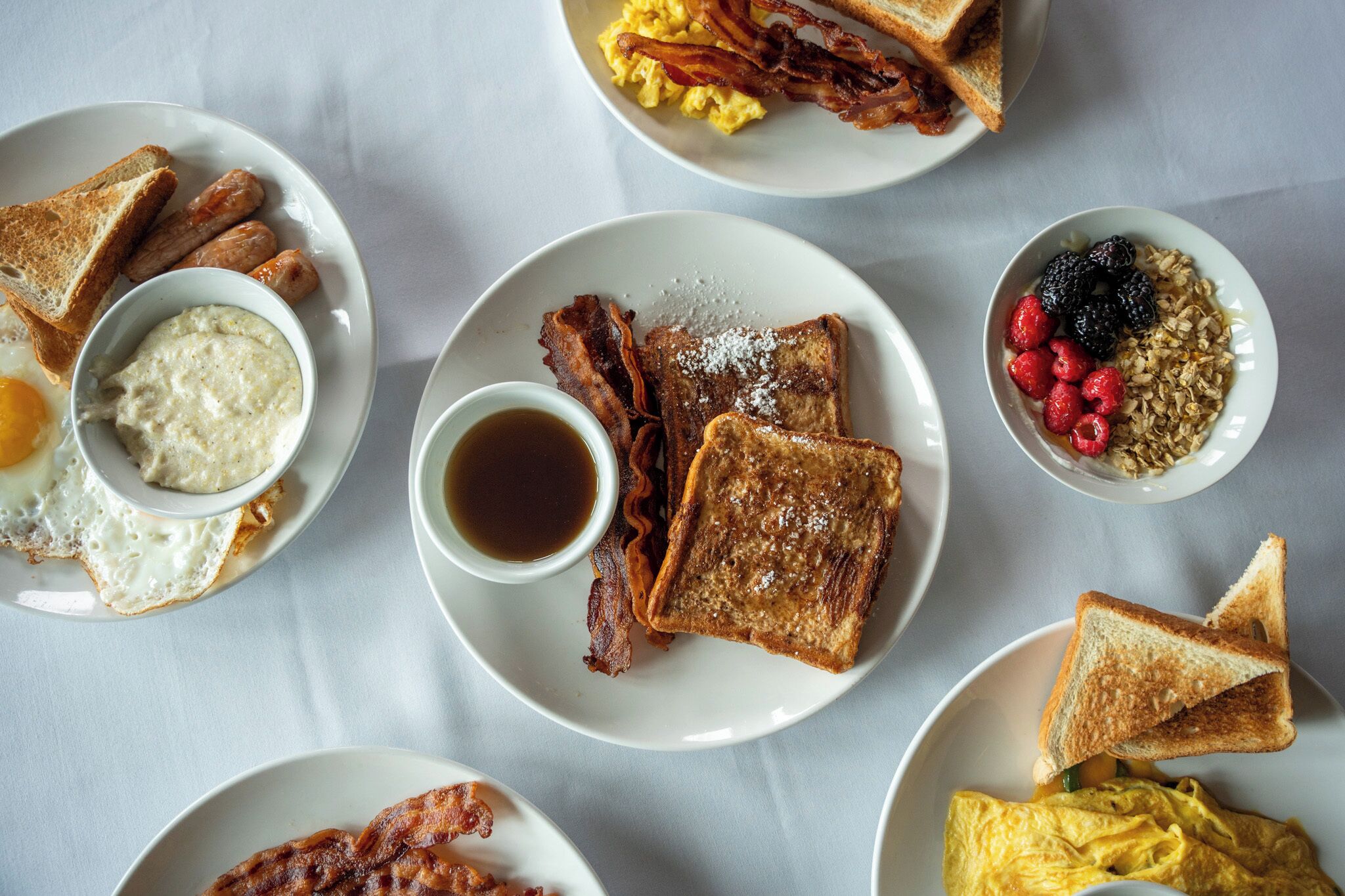 Un assortiment de mets pour le petit-déjeuner, comprenant des toasts, des œufs, du bacon, des saucisses et des fruits, le tout présenté sur des assiettes.