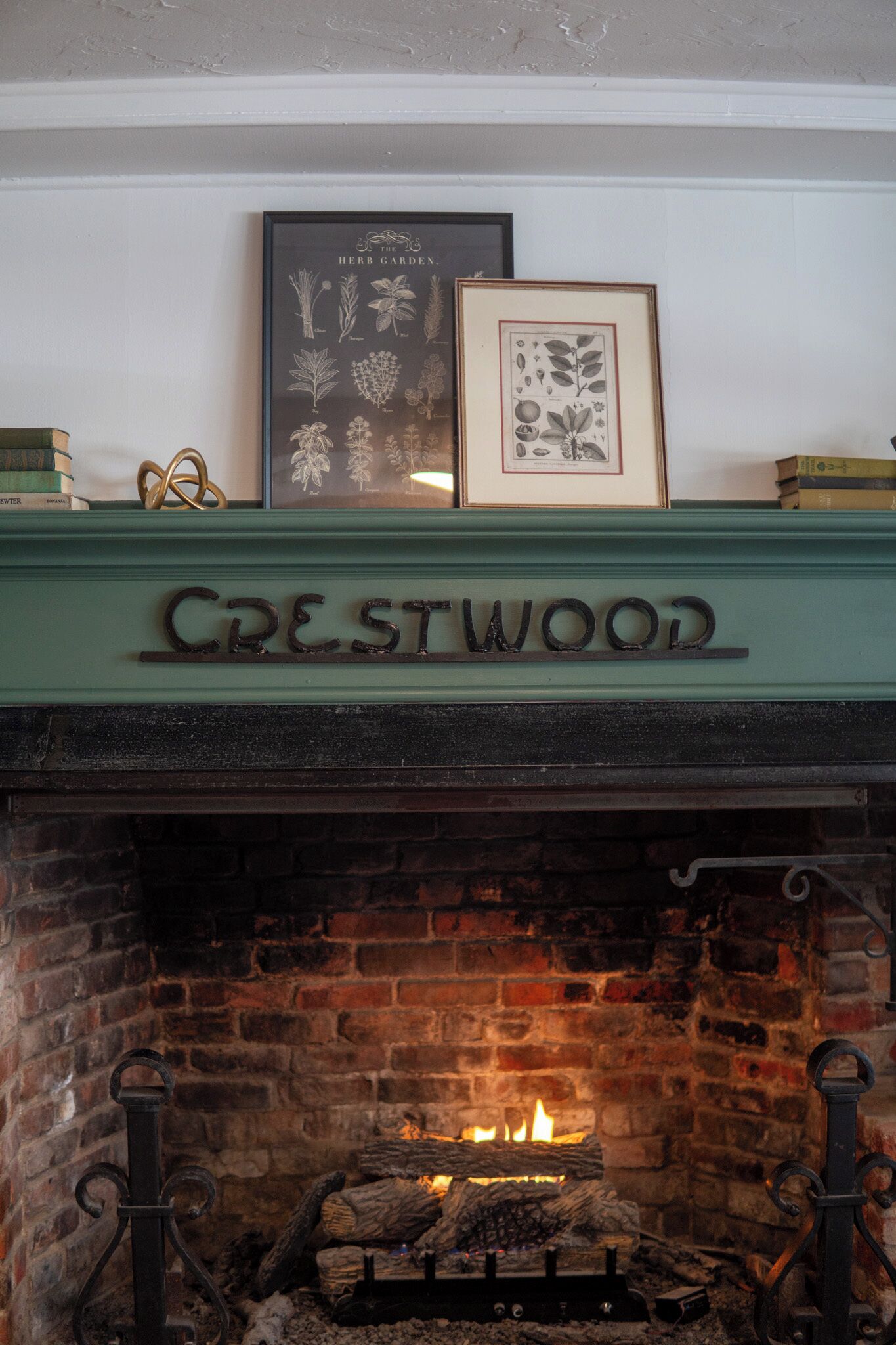 A fireplace with a 'Crestwood' sign above it, framed botanical prints, and a brick wall.