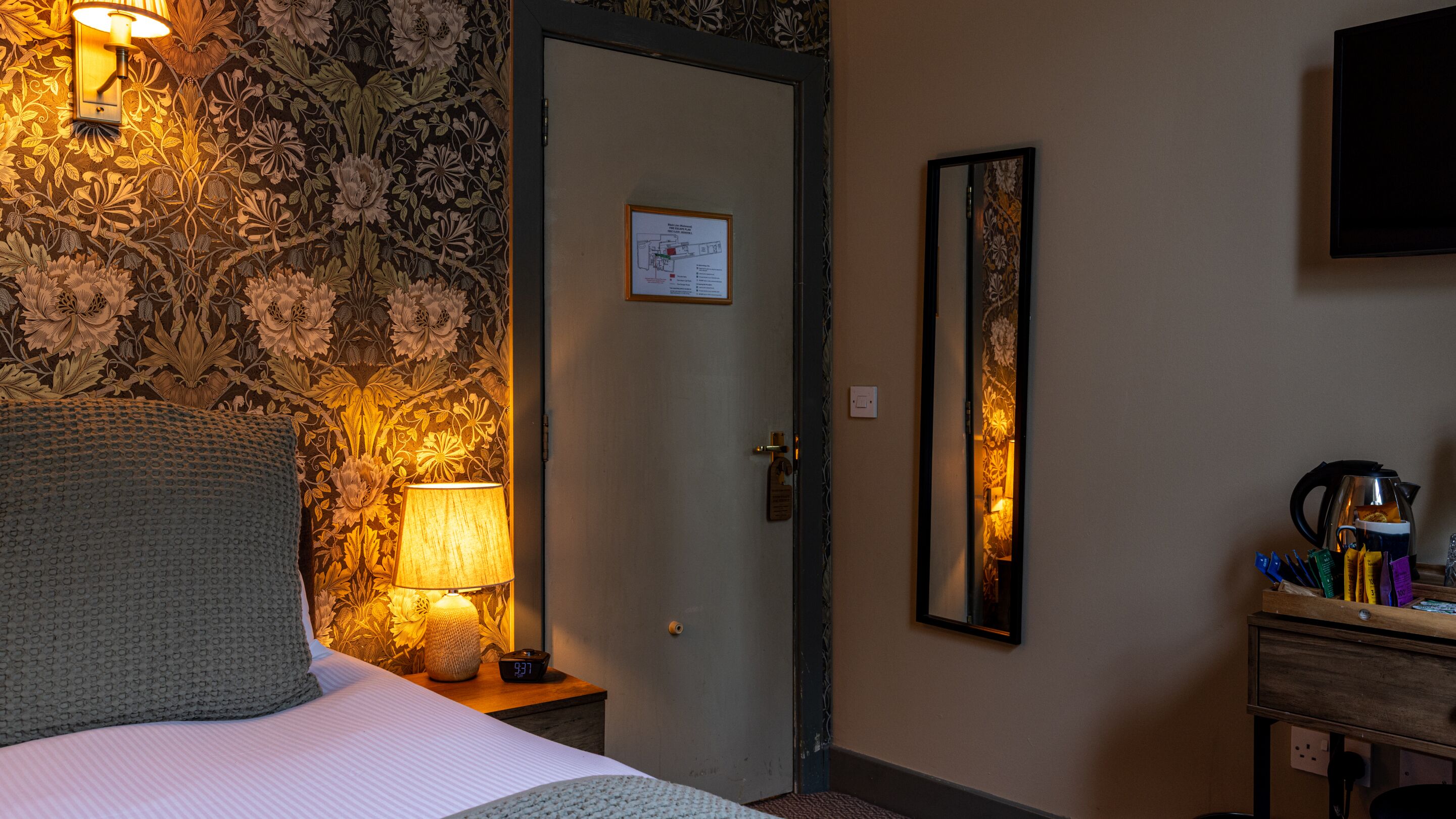 A hotel room with a bed, a nightstand with a lamp, a wall clock, a door with a map, and a mirror.