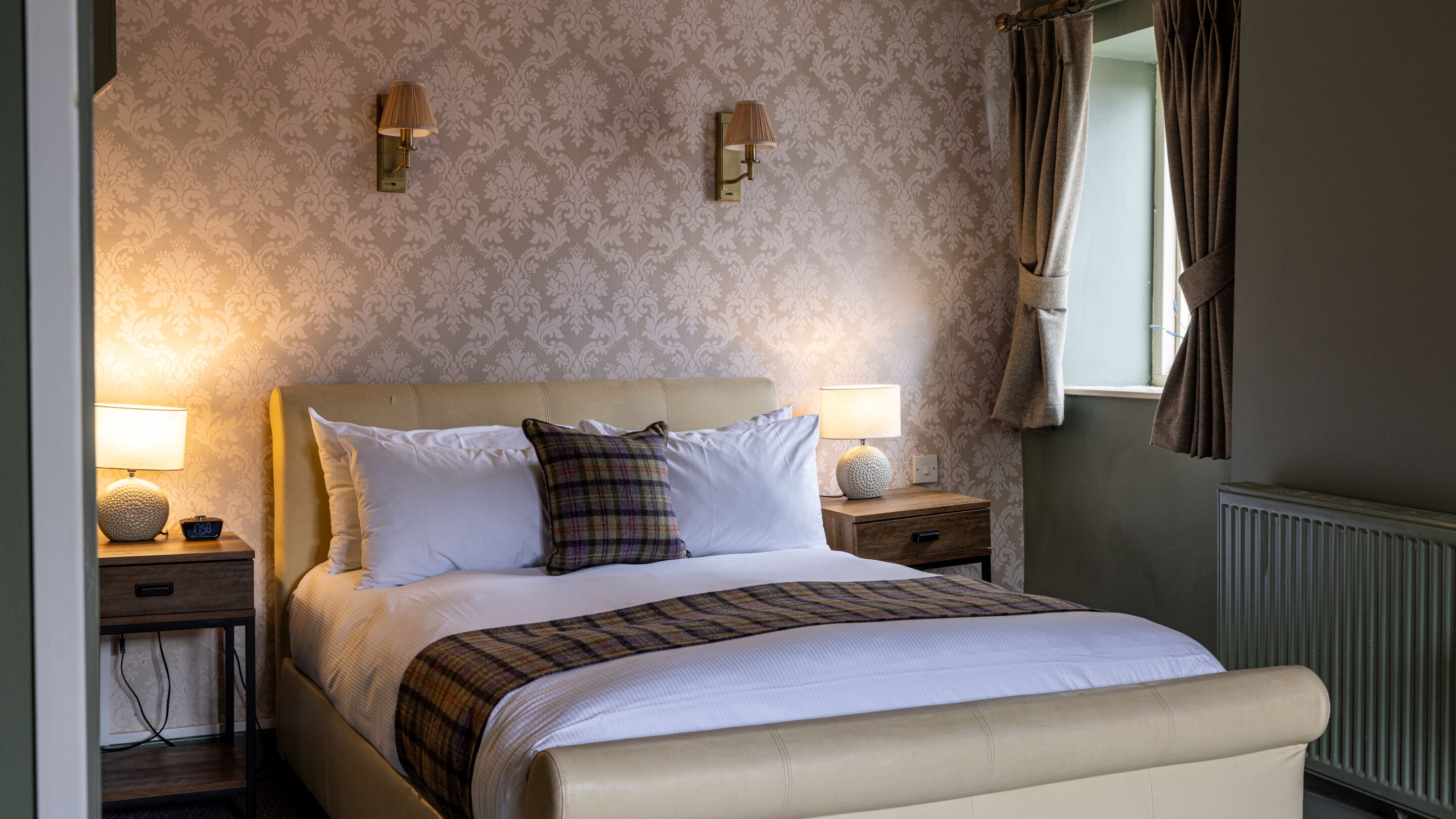 A neatly made bed with a headboard, two bedside lamps, a clock, and a window with curtains.