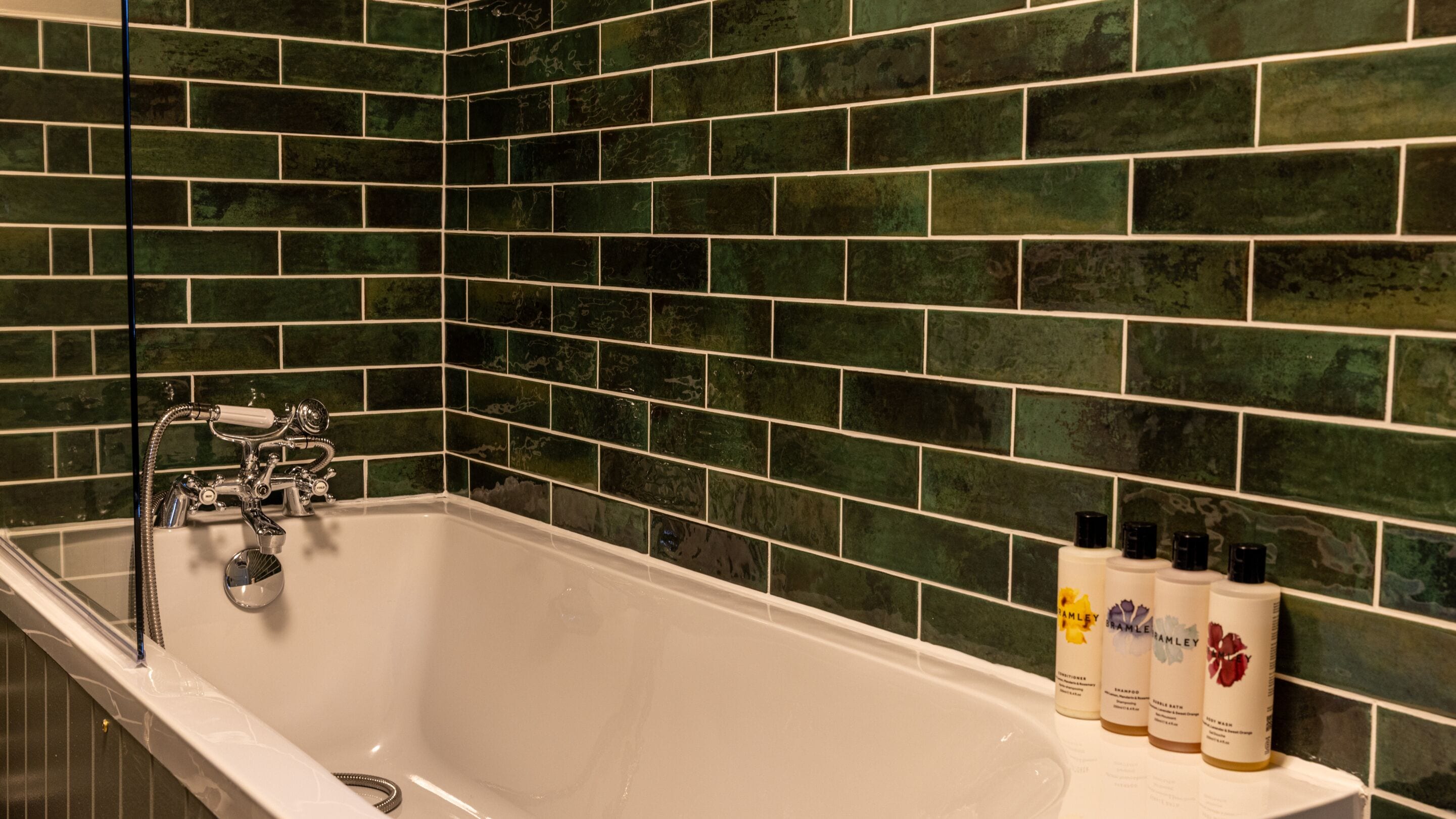 Un baño con una bañera blanca, paredes cubiertas de azulejos verdes y tres botellas de champú en el borde.