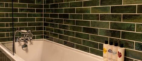 Un baño con una bañera blanca, paredes cubiertas de azulejos verdes y tres botellas de champú en el borde.