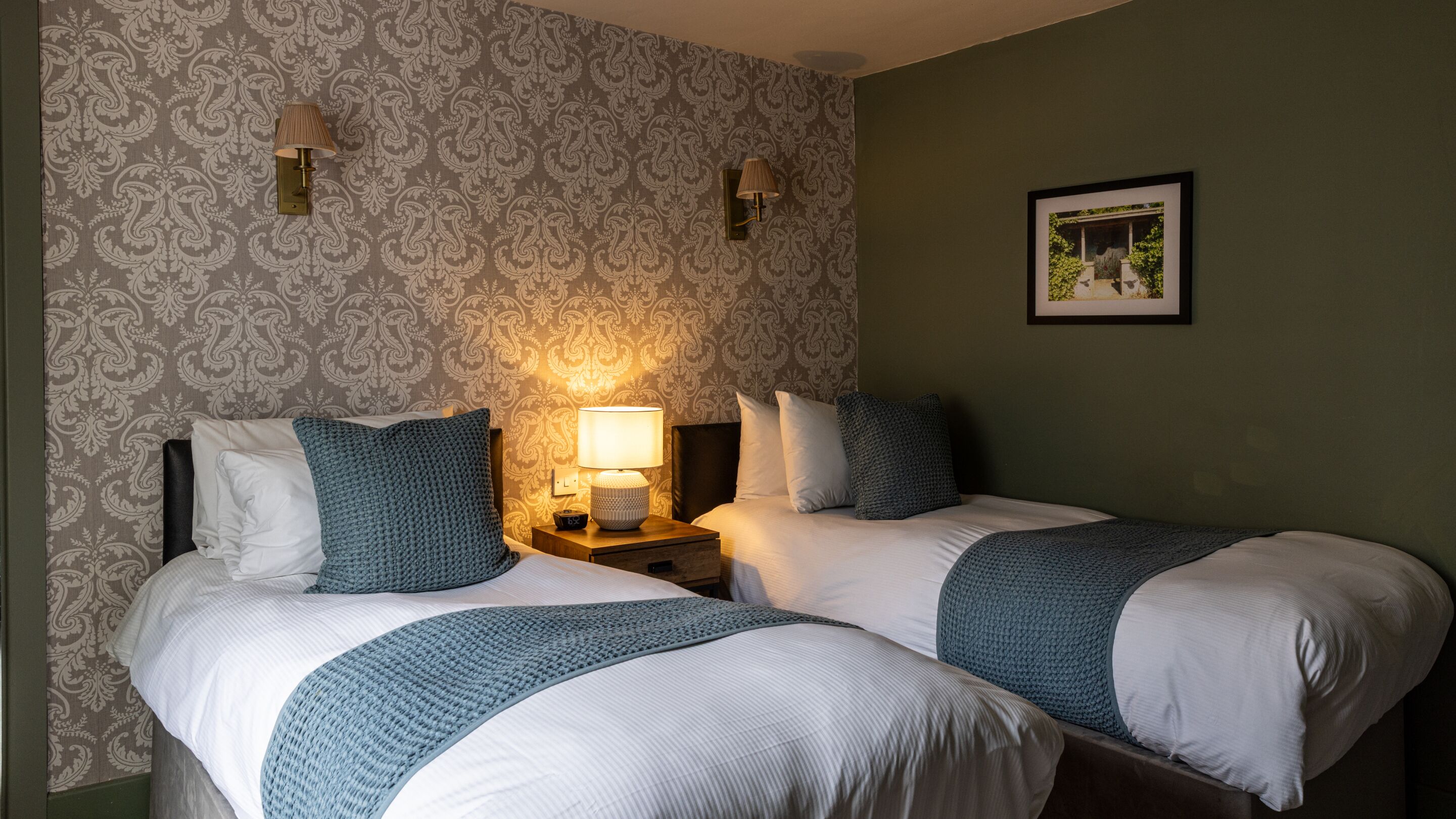 A hotel room with two beds, a nightstand with a lamp, patterned wallpaper, and a framed picture on the wall.