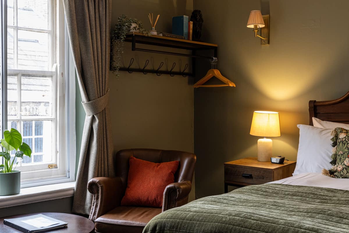 Hotelkamer met een bed, een stoel, een nachtkastje, een lamp, een raam met gordijnen, en een wandplank met boeken en een plant.