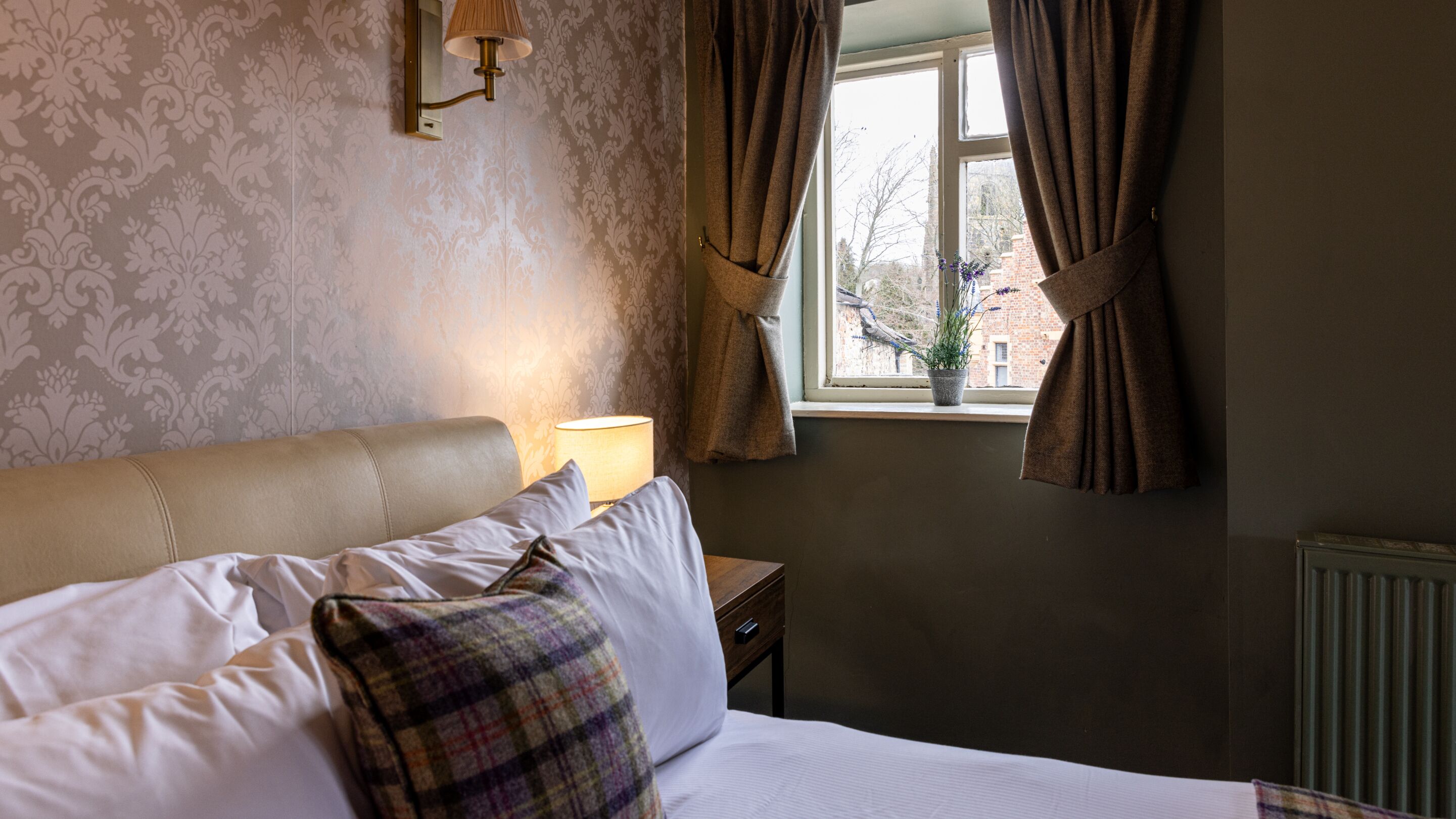 A bedroom with a bed, a nightstand, a lamp, a window with curtains, and a view of the outside.