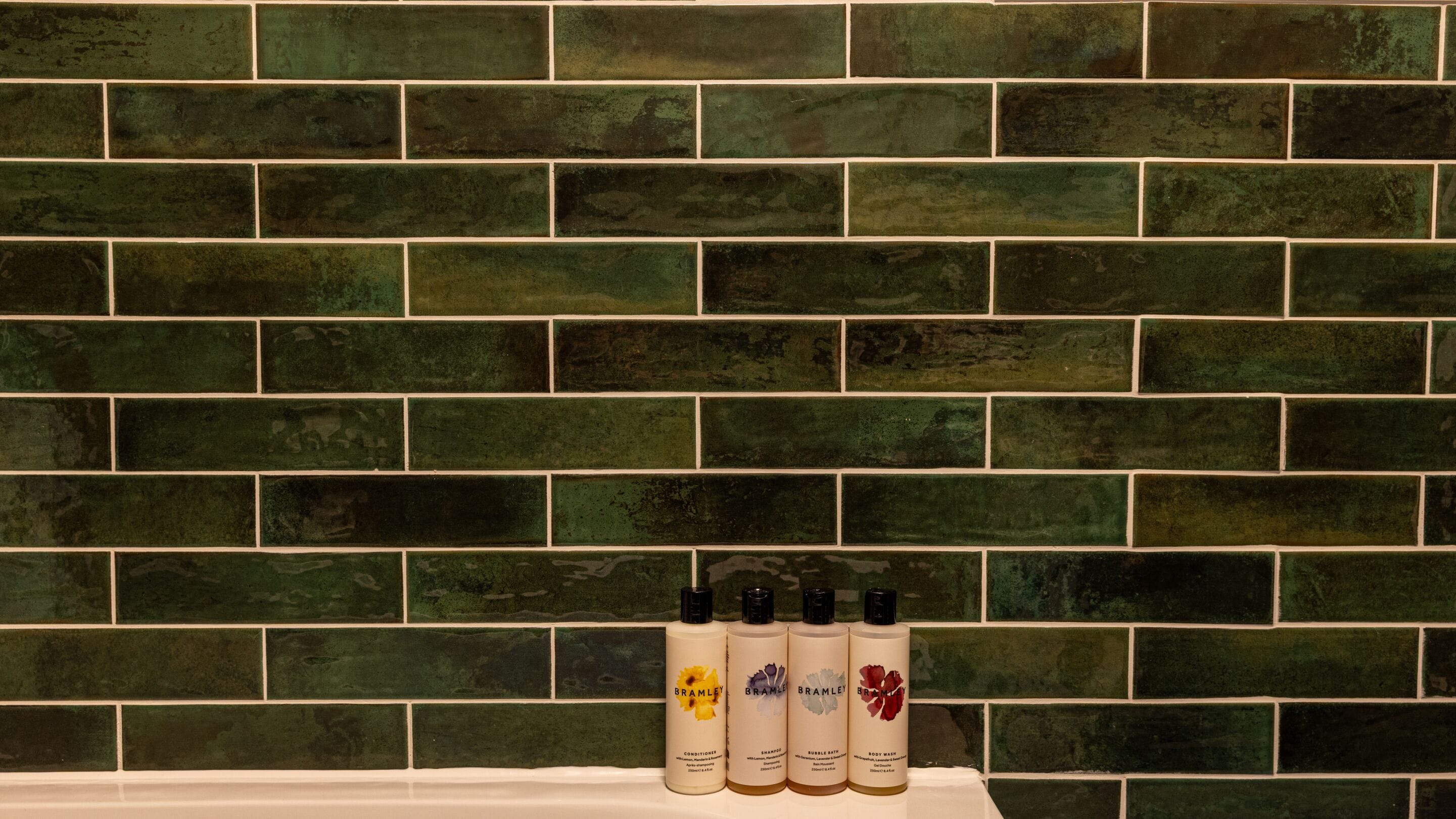 A bathroom with a green brick wall and four bottles of Bramble body wash on a counter.