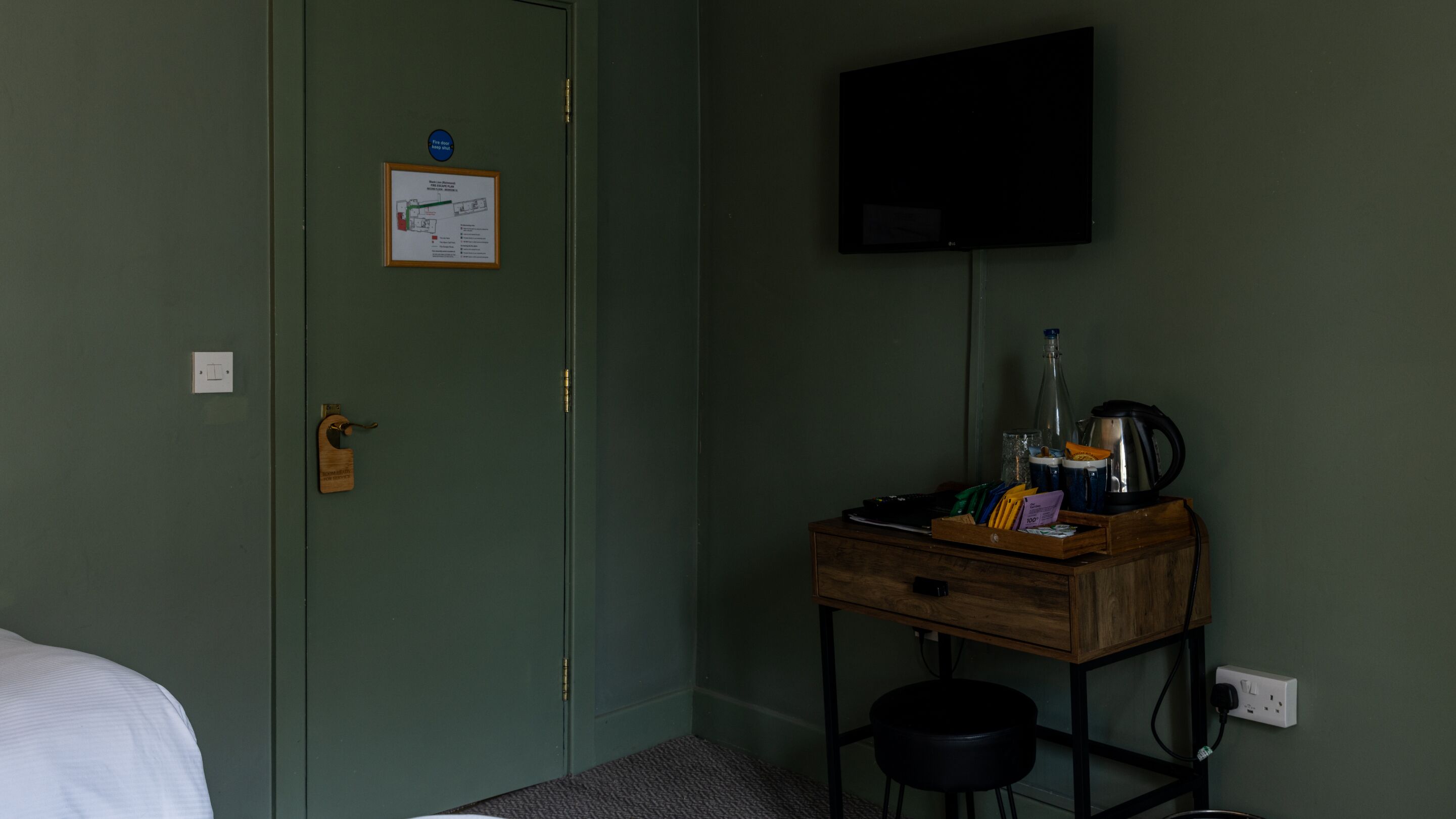 A room with a green door, a mounted TV, a wooden desk with a kettle, and a small stool.