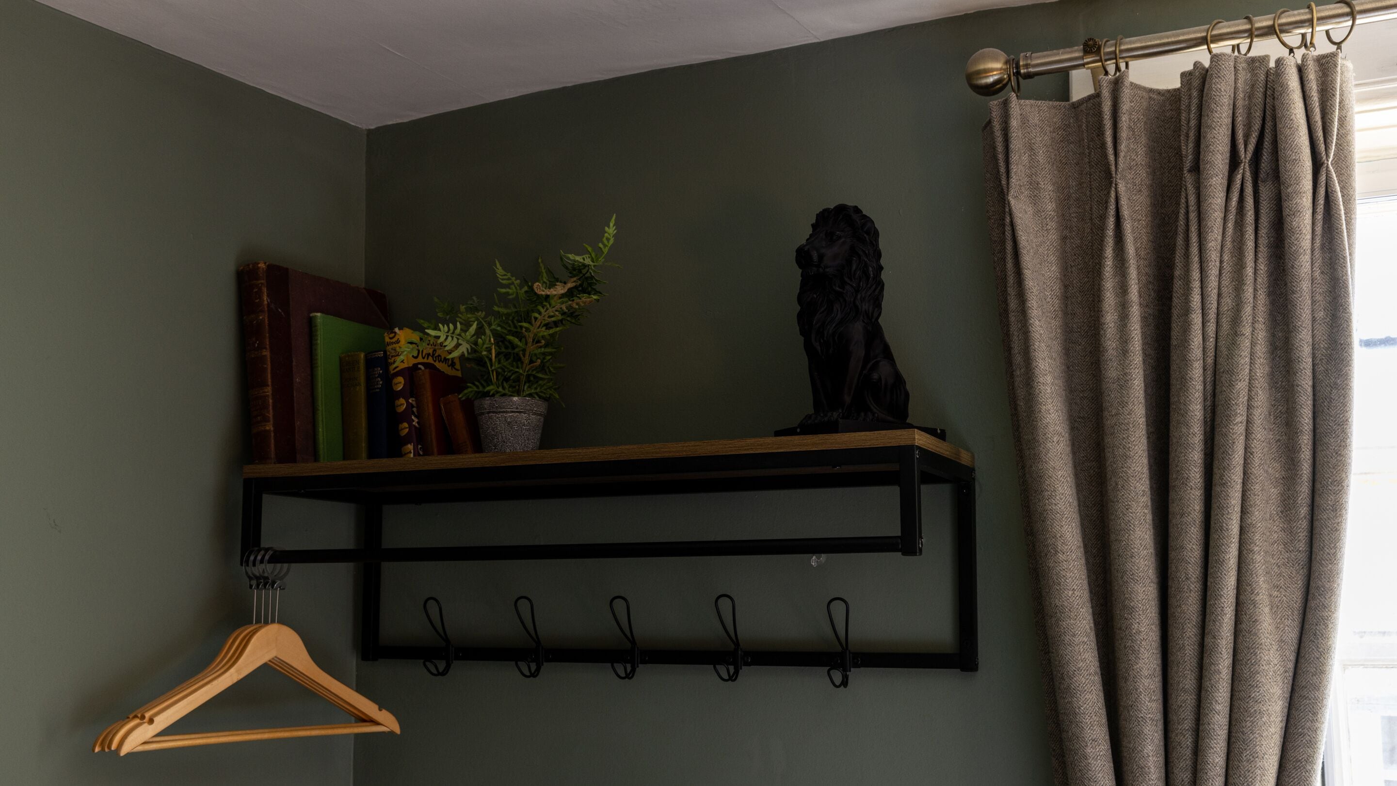 Una habitación con una pared verde, un estante negro con libros y una planta, una estatua negra, un perchero de madera y una ventana con cortinas.