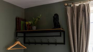 Una habitación con una pared verde, un estante negro con libros y una planta, una estatua negra, un perchero de madera y una ventana con cortinas.