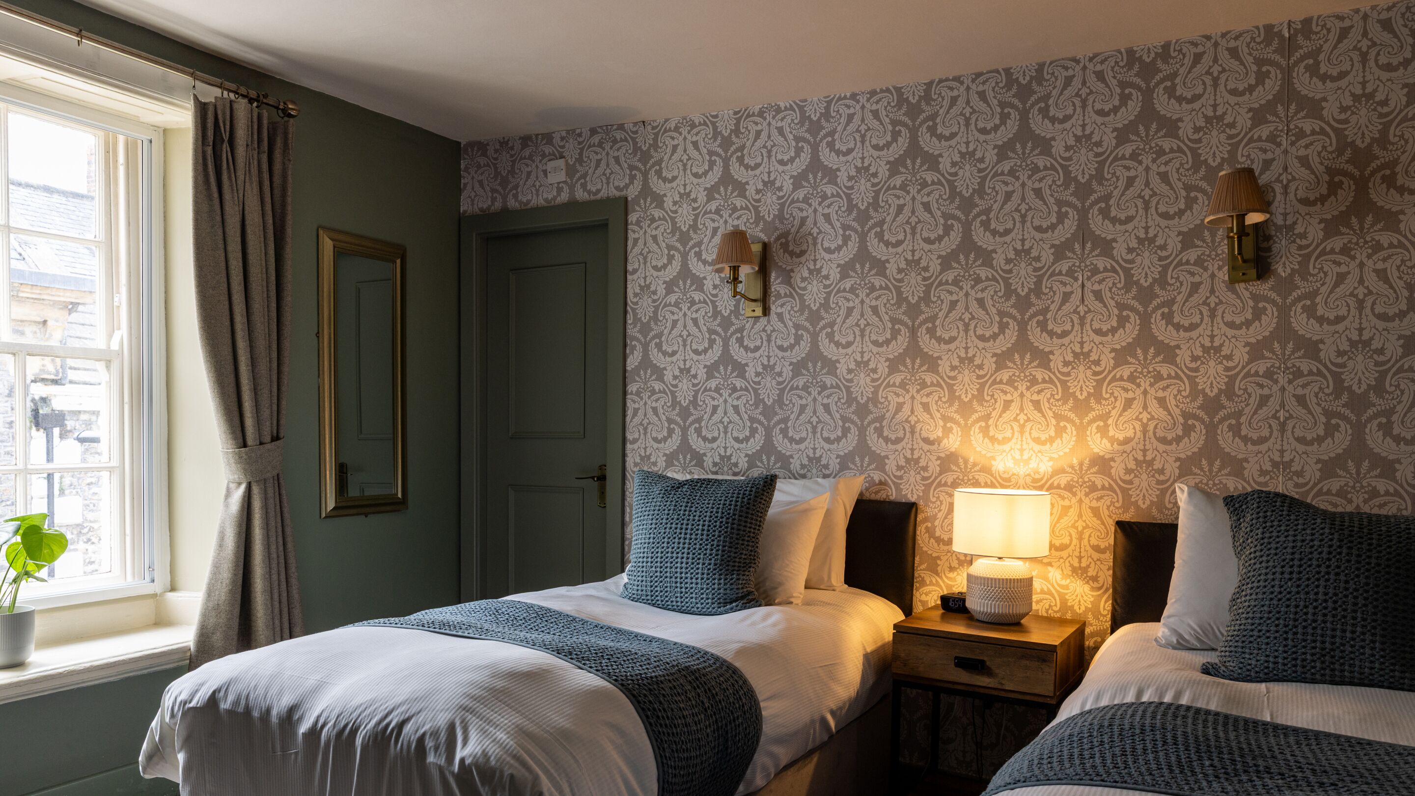 Une chambre avec deux lits, une fenêtre avec des rideaux, un mur recouvert d’un papier peint à motifs et deux lampes fixées au mur.