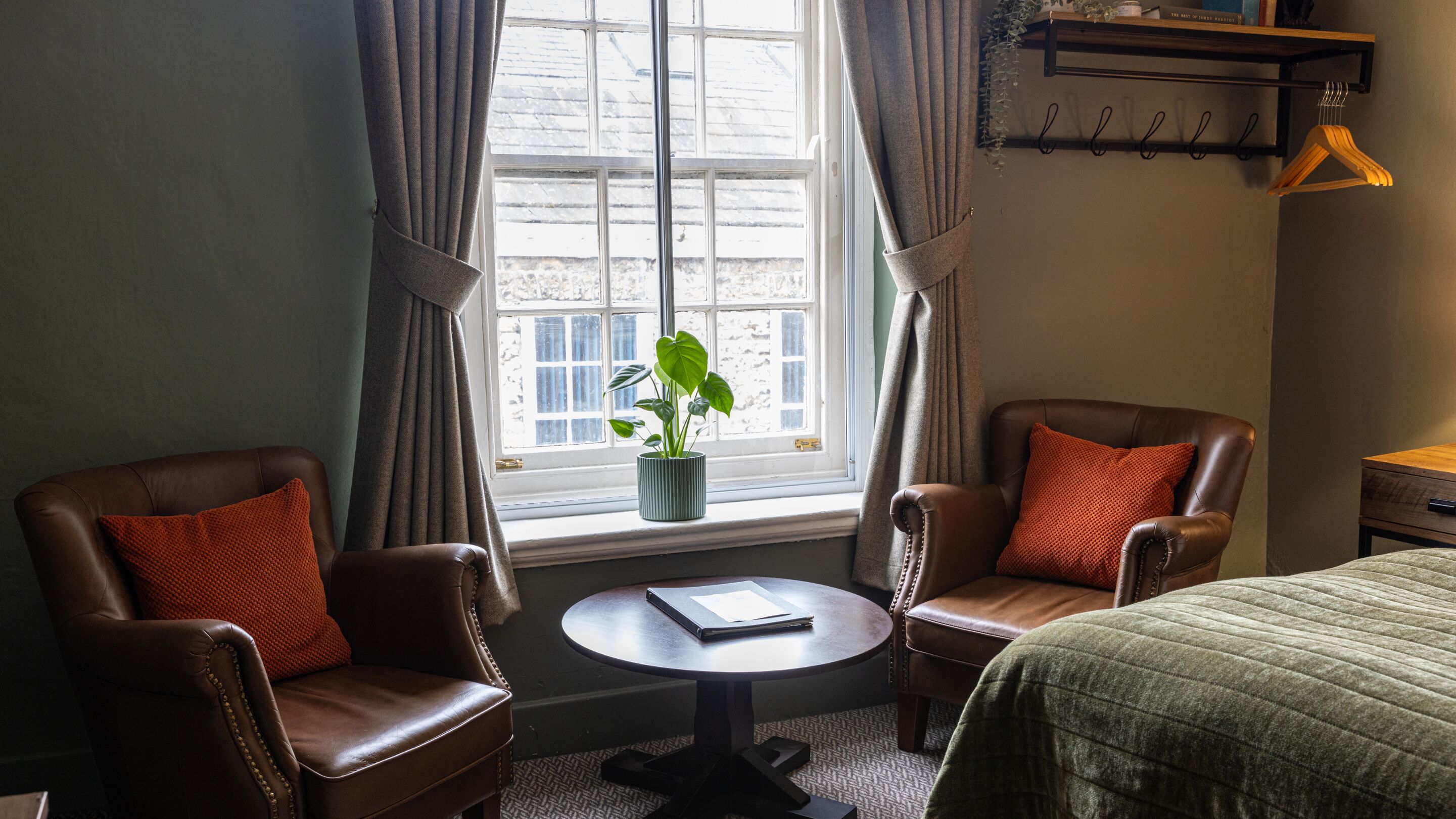A hotel room with two leather armchairs, a round table, a bed, a window with curtains, a potted plant, and a wooden shelf with hangers.