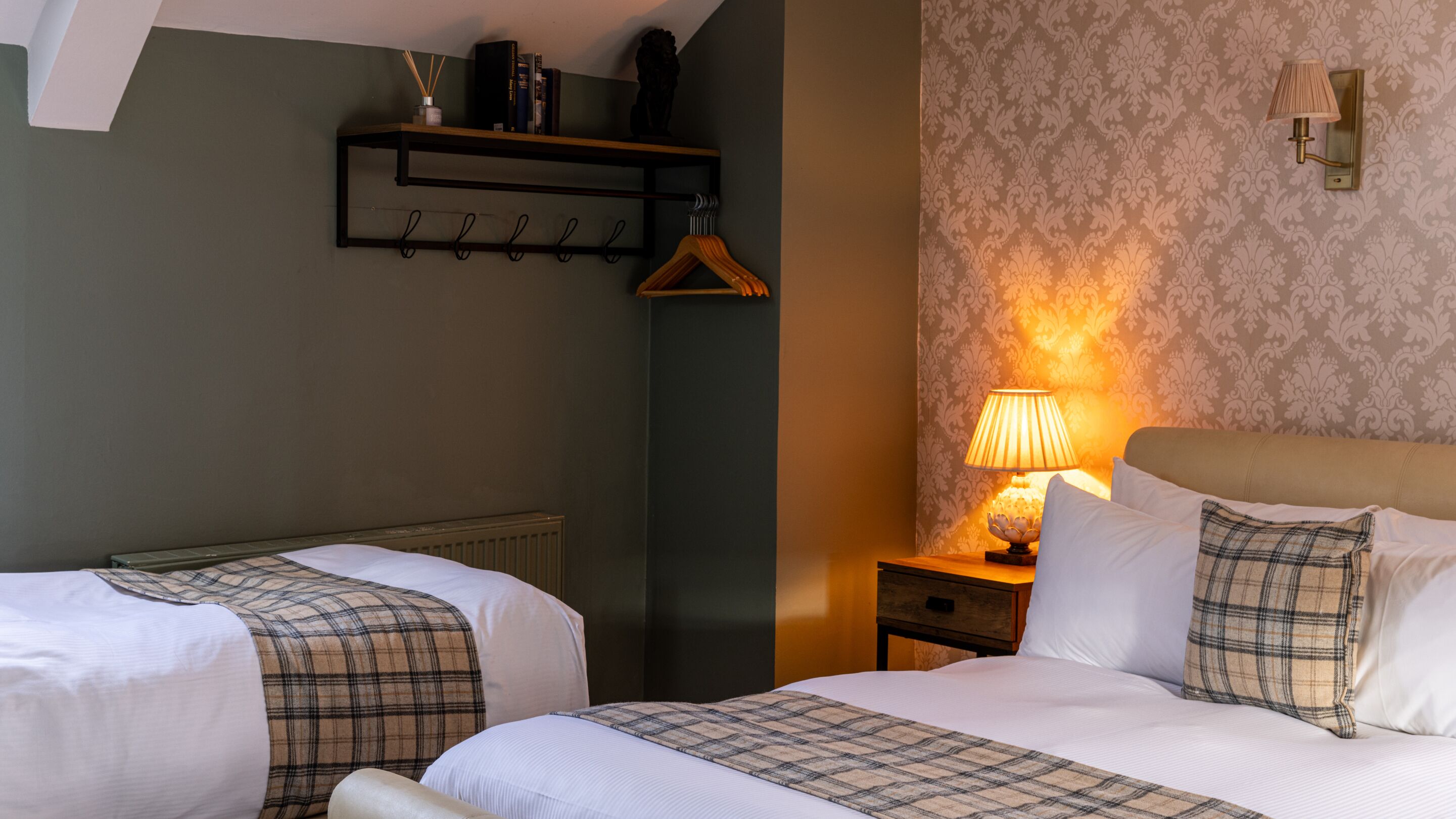 A hotel room with two beds, a nightstand with a lamp, a shelf with books and a vase, and a patterned wallpaper.