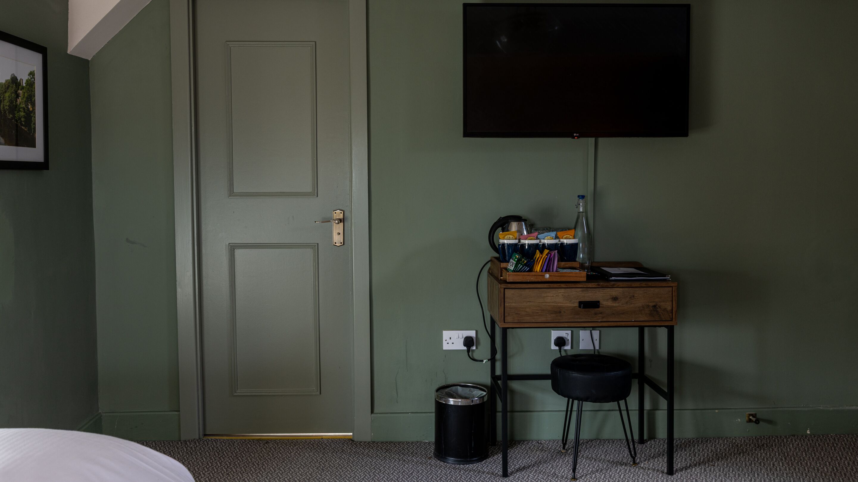 A room with a closed door, a desk with a flat-screen TV, a trash can, and a power outlet.