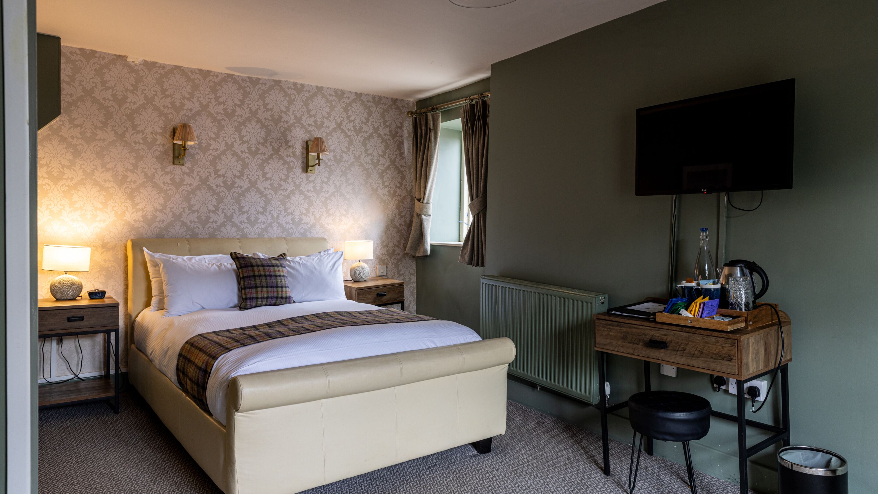 A hotel room with a bed, bedside tables, a desk with a chair, a radiator, a TV, and a window with curtains.