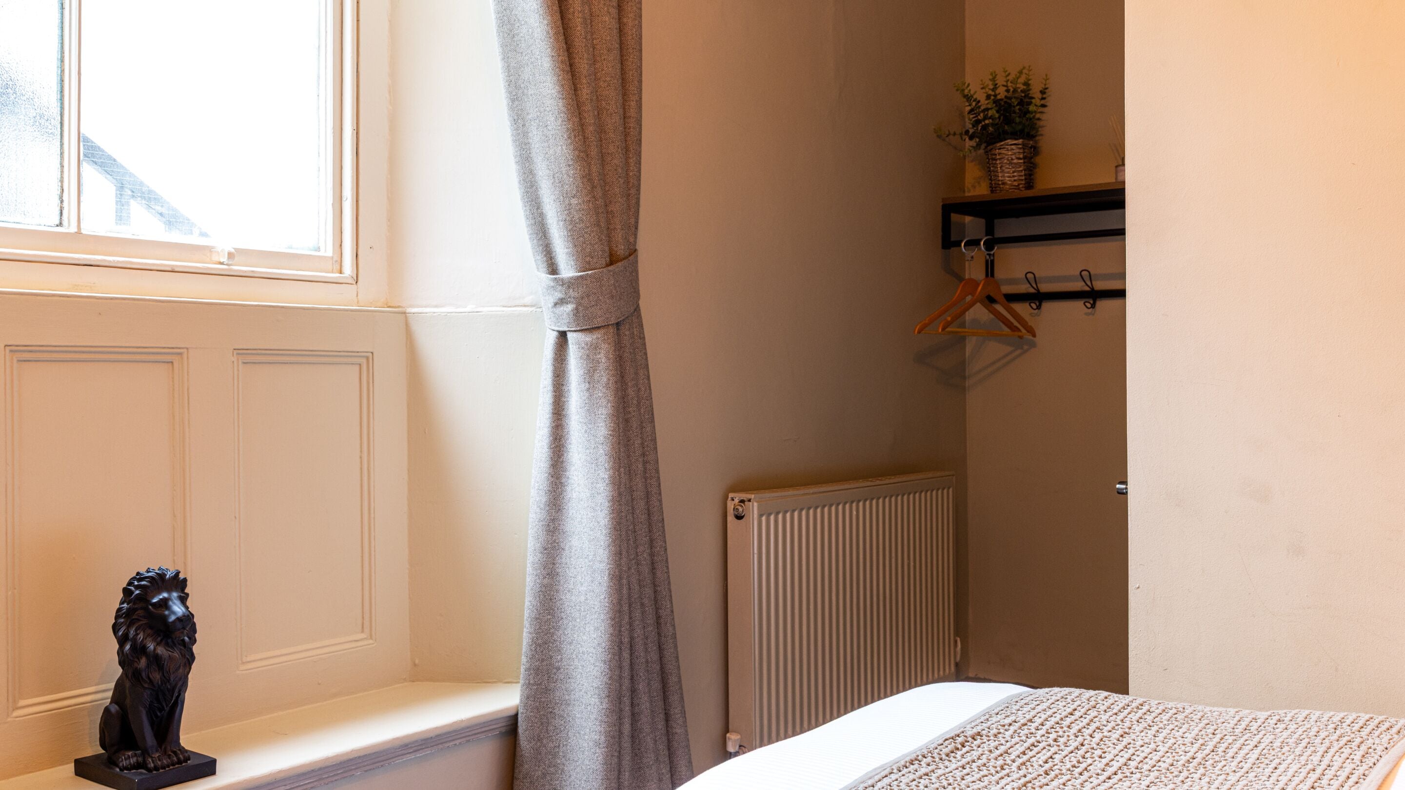 A bedroom with a radiator, a window with a curtain, a wooden shelf with a plant, and a sculpture on the windowsill.