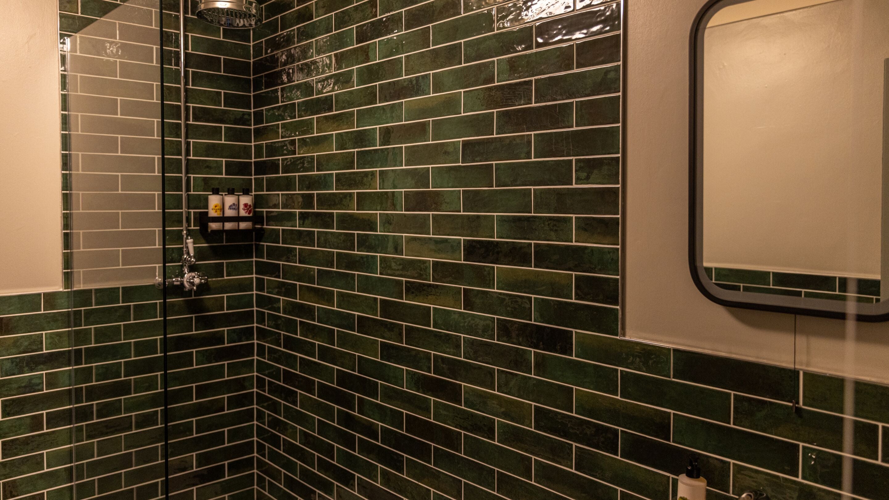 Rincón de un baño con baldosas de mosaico verde, un box de ducha de vidrio y un pequeño estante con artículos de aseo.
