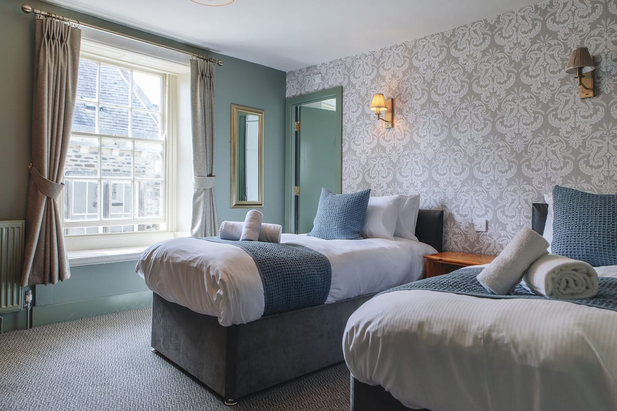 Een kamer met twee bedden, een raam met gordijnen, een spiegel en behang met een patroon.