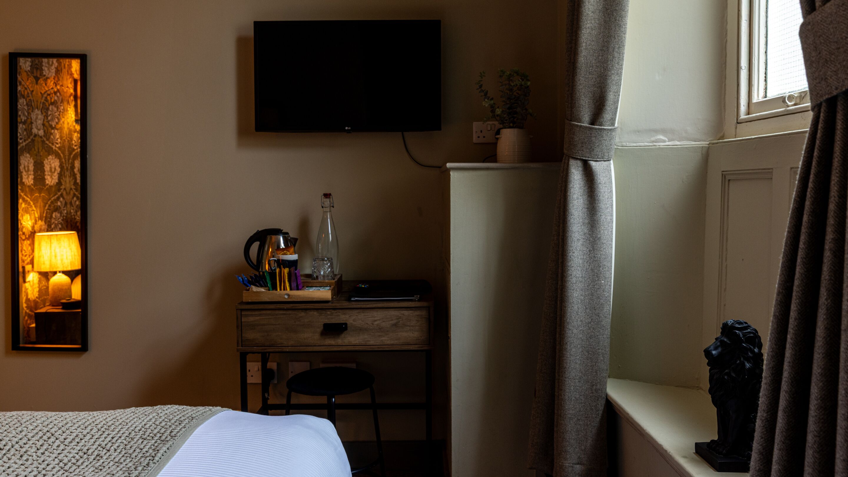 Habitación de hotel con cama, escritorio con computadora, televisor montado en la pared, ventana con cortinas y una estatua en un estante.