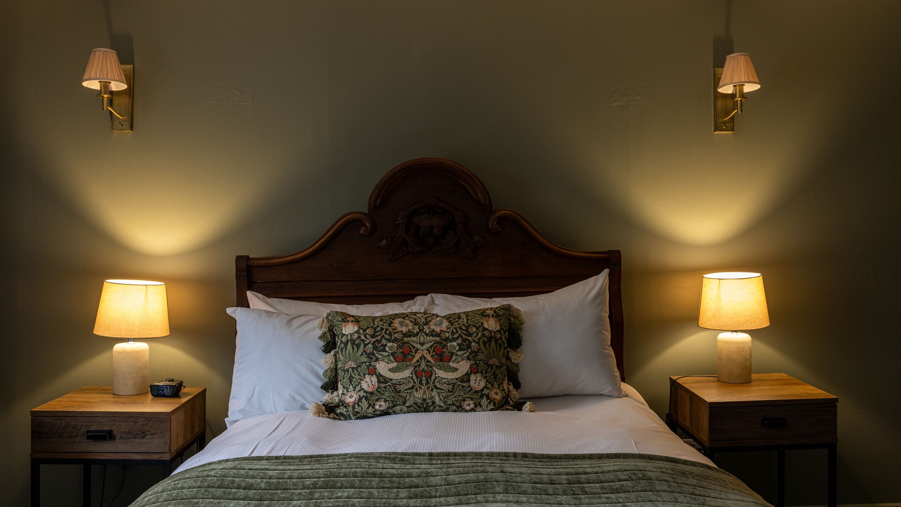 A bed with a wooden headboard, two bedside lamps, a nightstand with a clock, and a floral pillow.