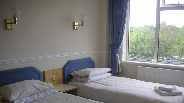 A hotel room with two single beds, a nightstand, a window with a view of trees, and wall-mounted lamps. - The Bridge Hotel (Scunthorpe)