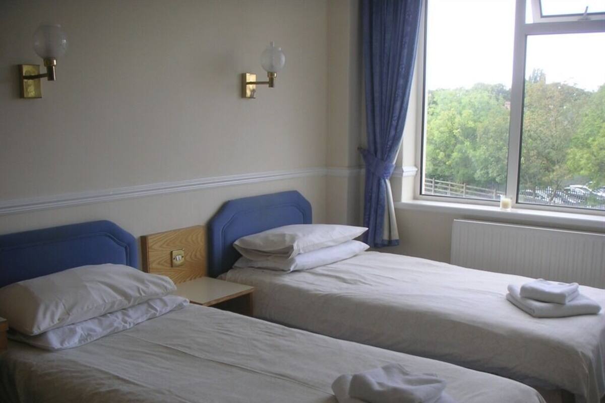 A hotel room with two single beds, a nightstand, a window with a view of trees, and wall-mounted lamps.