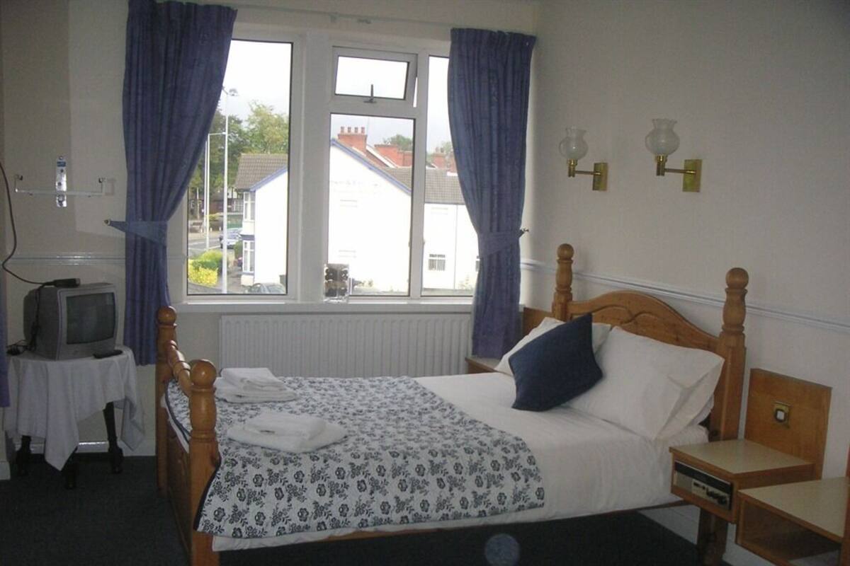 A bedroom with a wooden bed, a small bedside table, a nightstand, a television on a side table, and a window with curtains.