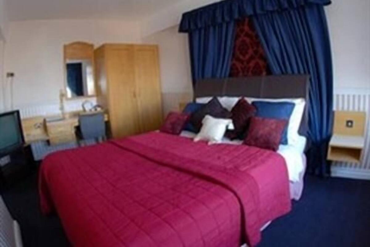 A hotel room with a large bed, a wooden wardrobe, a desk with a chair, and a window with curtains.