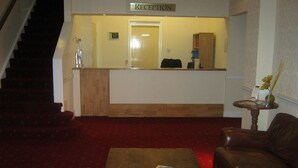 A reception area with a wooden reception desk, a staircase, a red carpeted floor, a brown leather sofa, and a small round table with a vase. - The Bridge Hotel (Scunthorpe)