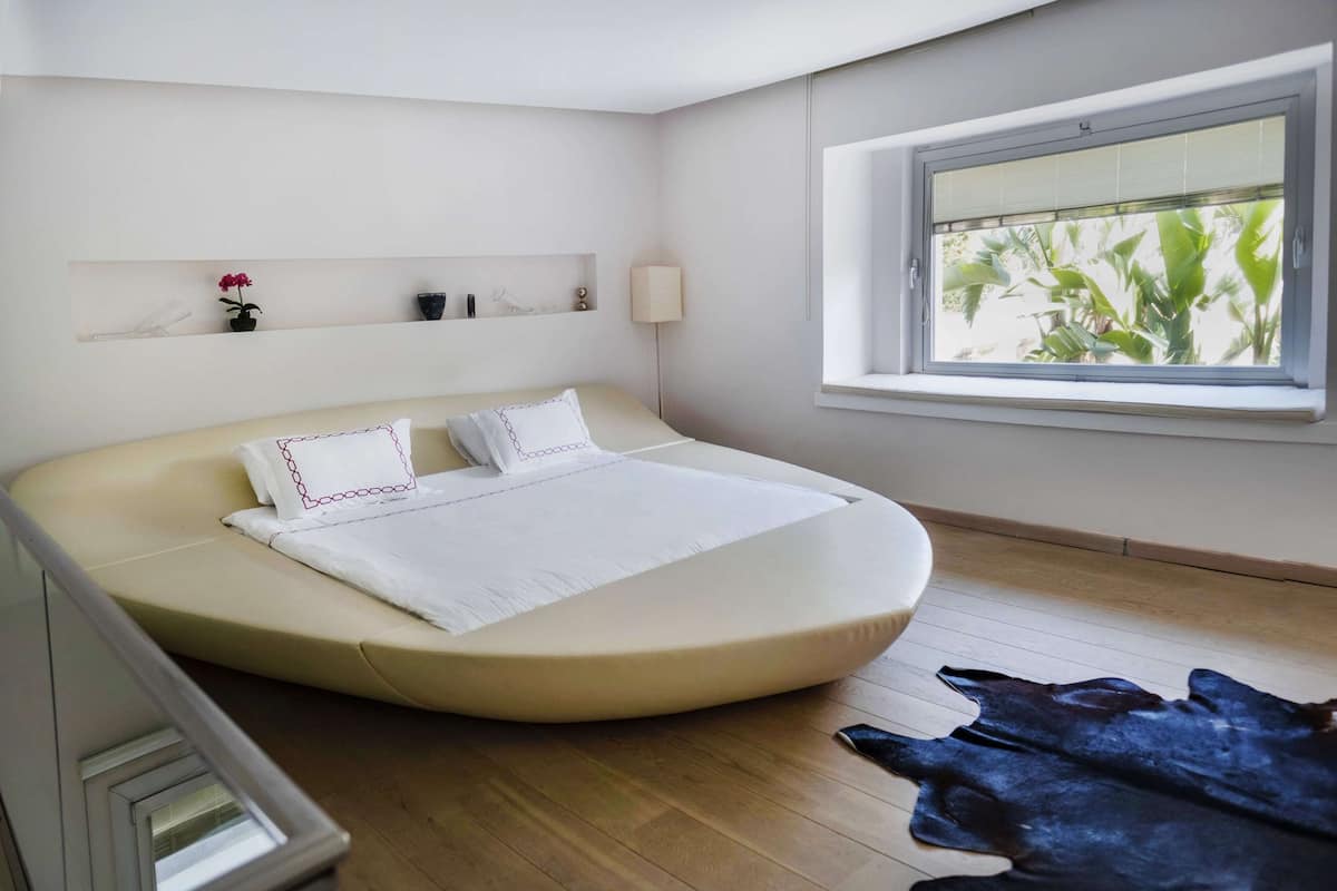 A modern bedroom with a curved bed, a window with a view of greenery, a bedside lamp, and a small shelf with decorative items.