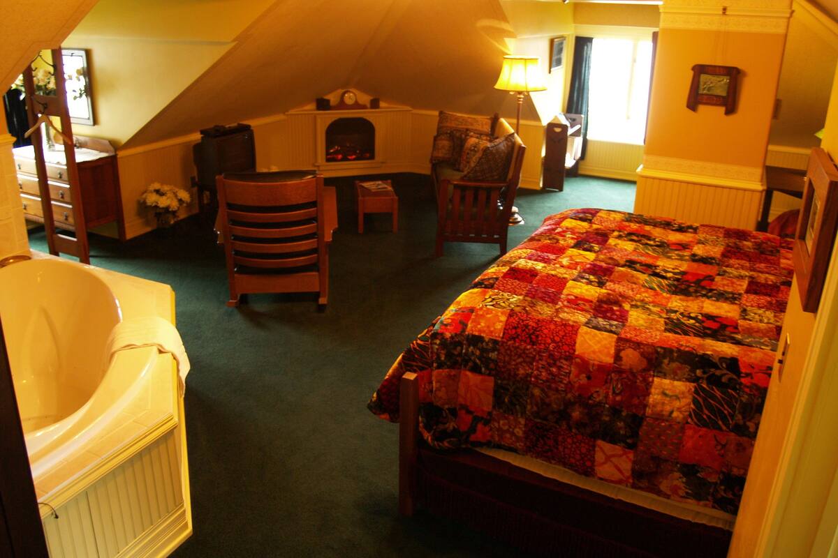 A bedroom with a large bed, a chair, a small table, and a fireplace.